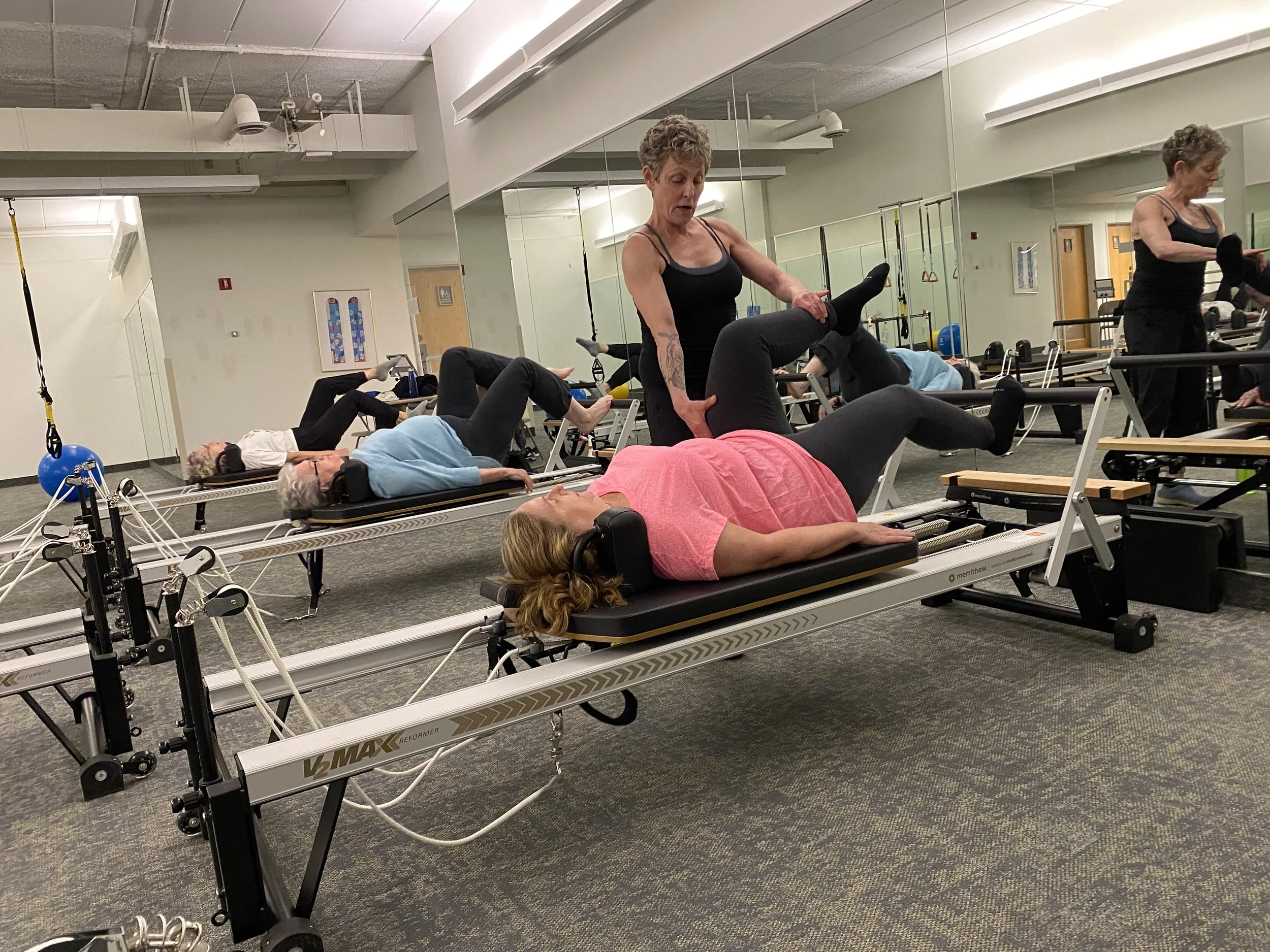 Group of elderly women doing Pilates exercises on reformer machines in a fitness studio, supervised by an instructor.