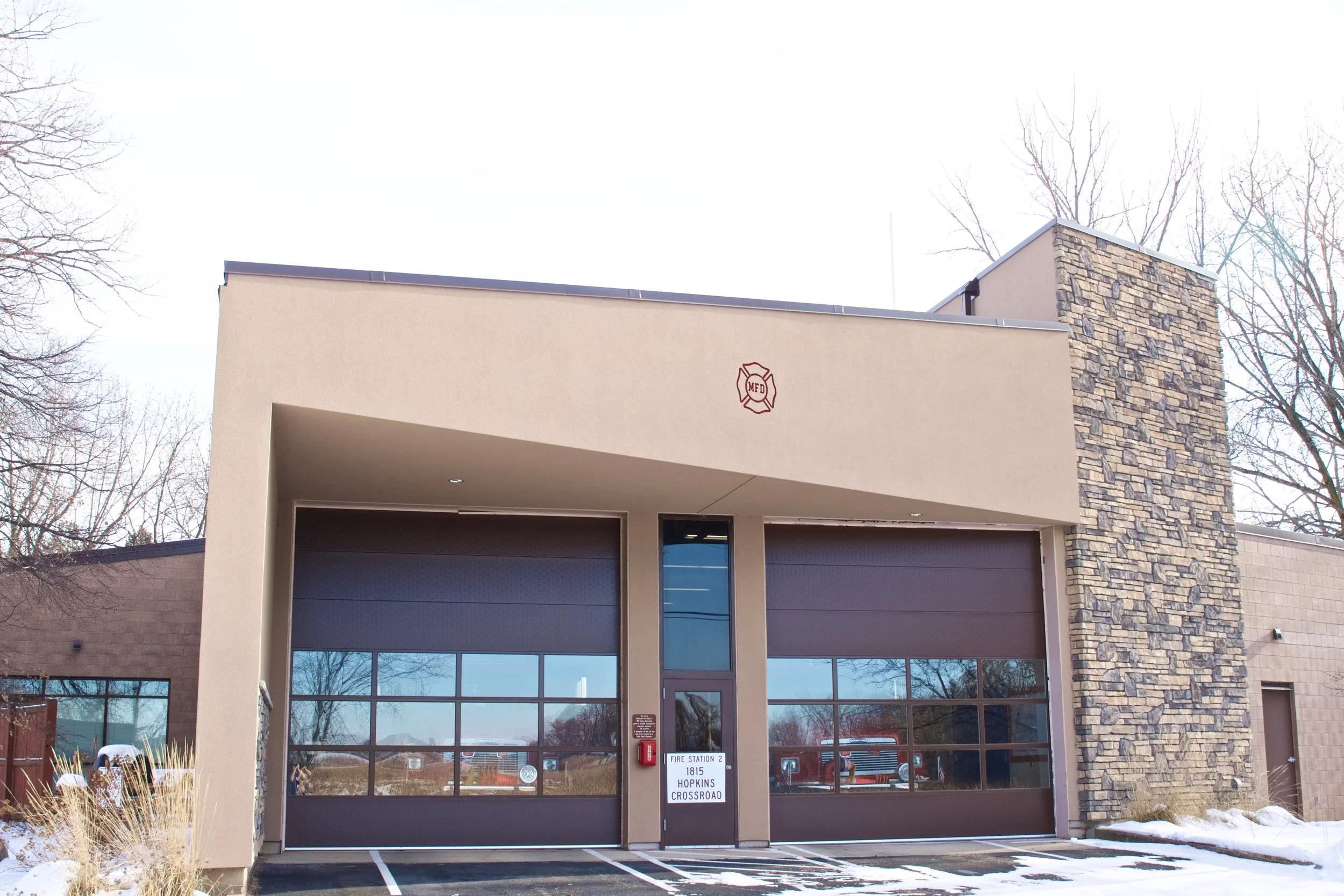 Modern fire station with two large garage doors, a sign reading 'Fire Station 2, 1815 Hopkins Crossroad', and a fire department emblem on the front wall. Snow on the ground and leafless trees in the background.
