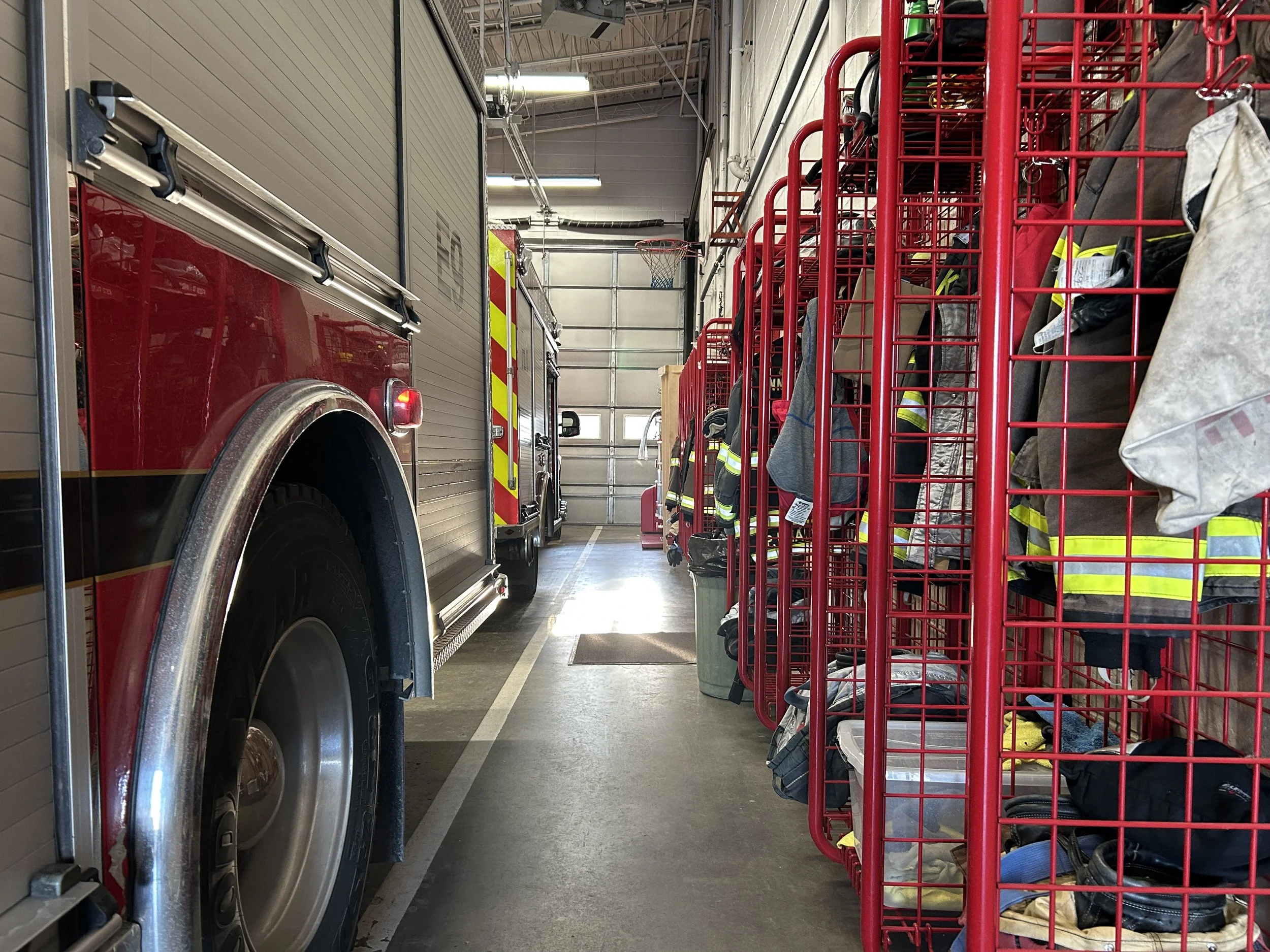 Firefighter gear and uniforms stored in red metal carts inside a fire station near a fire truck.