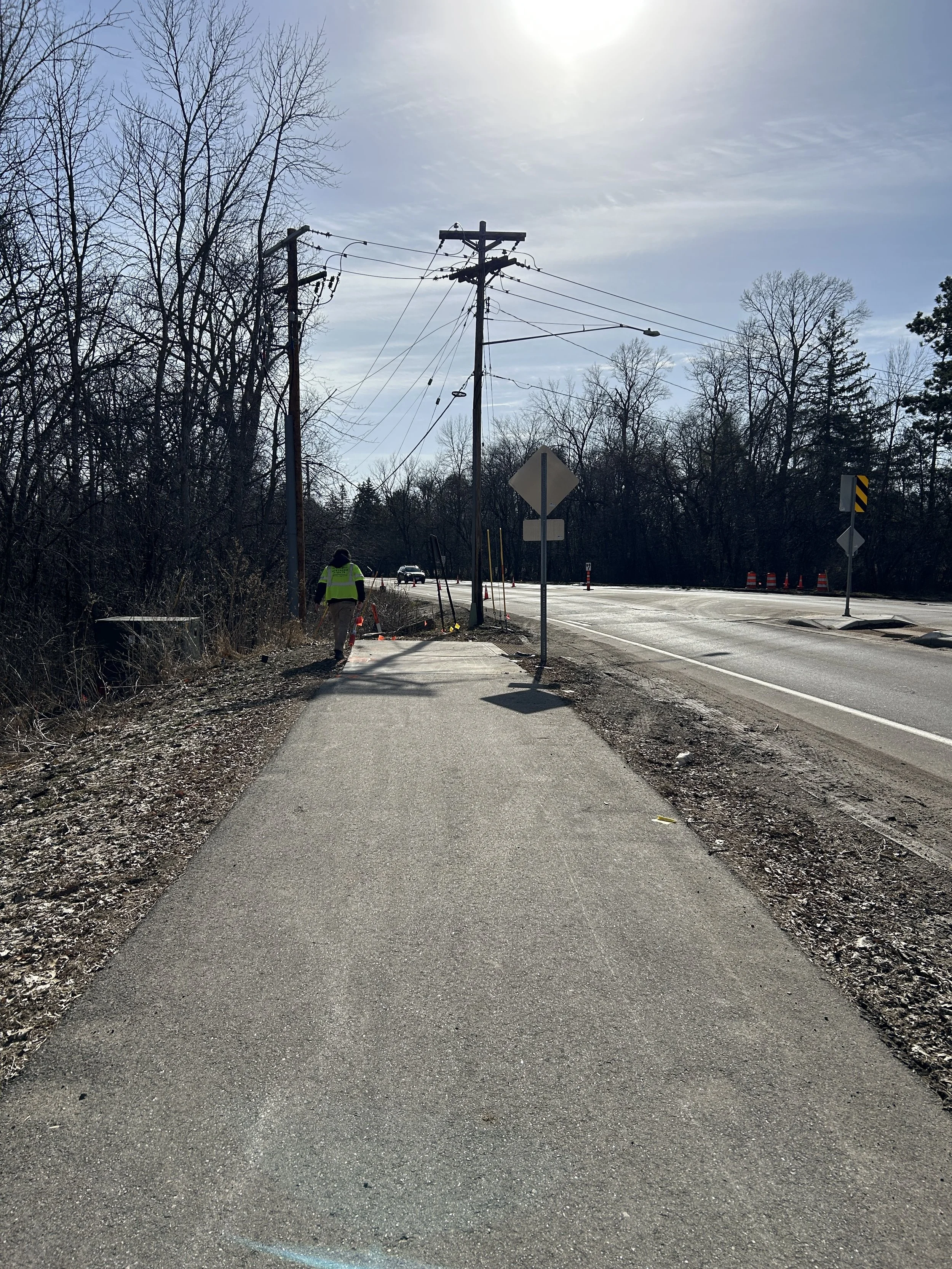 A sidewalk under construction with a worker in a yellow safety vest, near utility poles and trees, with a roadway in the background.