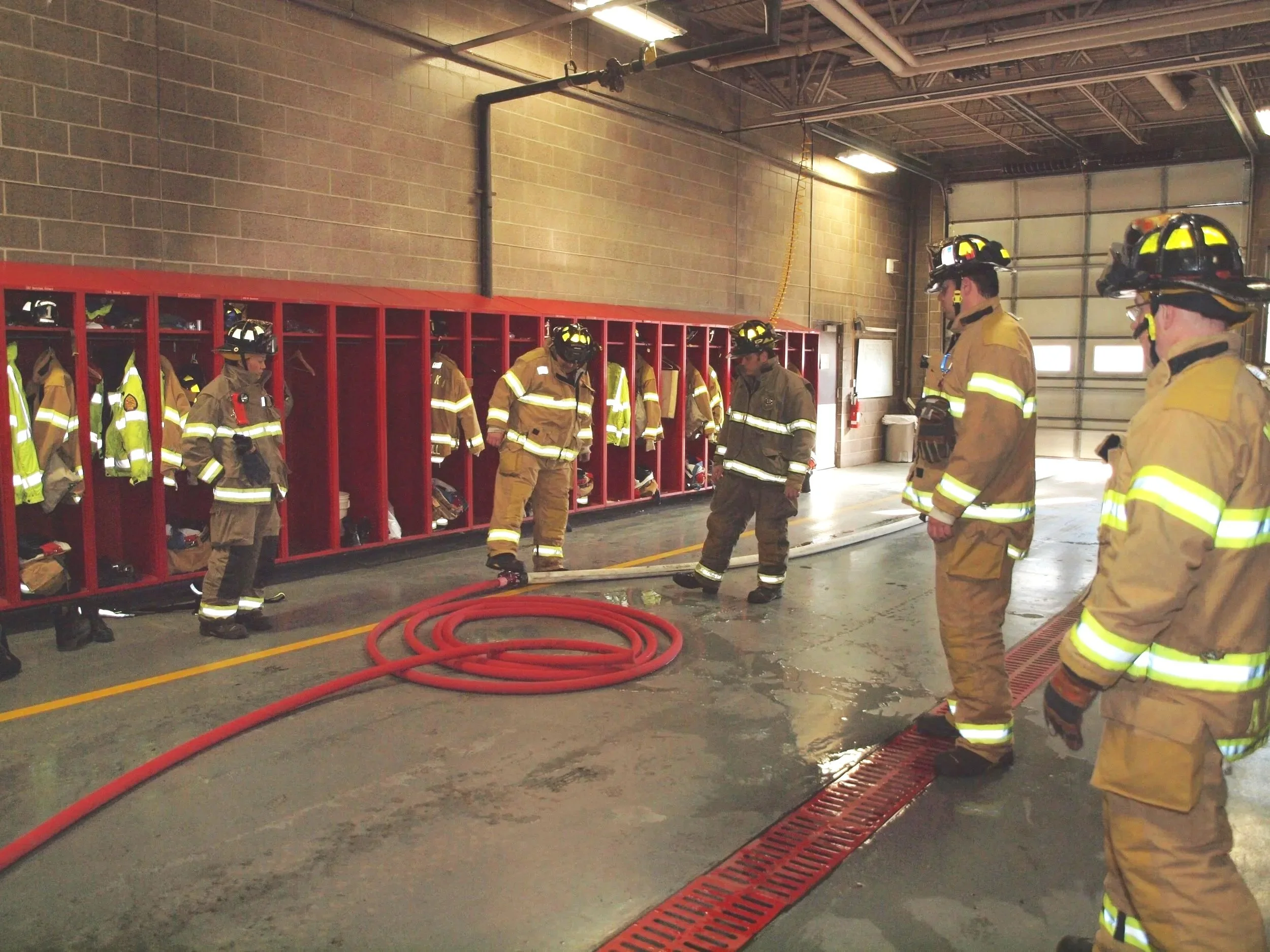 Group of firefighters in firefighting gear standing in a fire station, with red lockers containing helmets and other gear, and fire hoses on the ground.