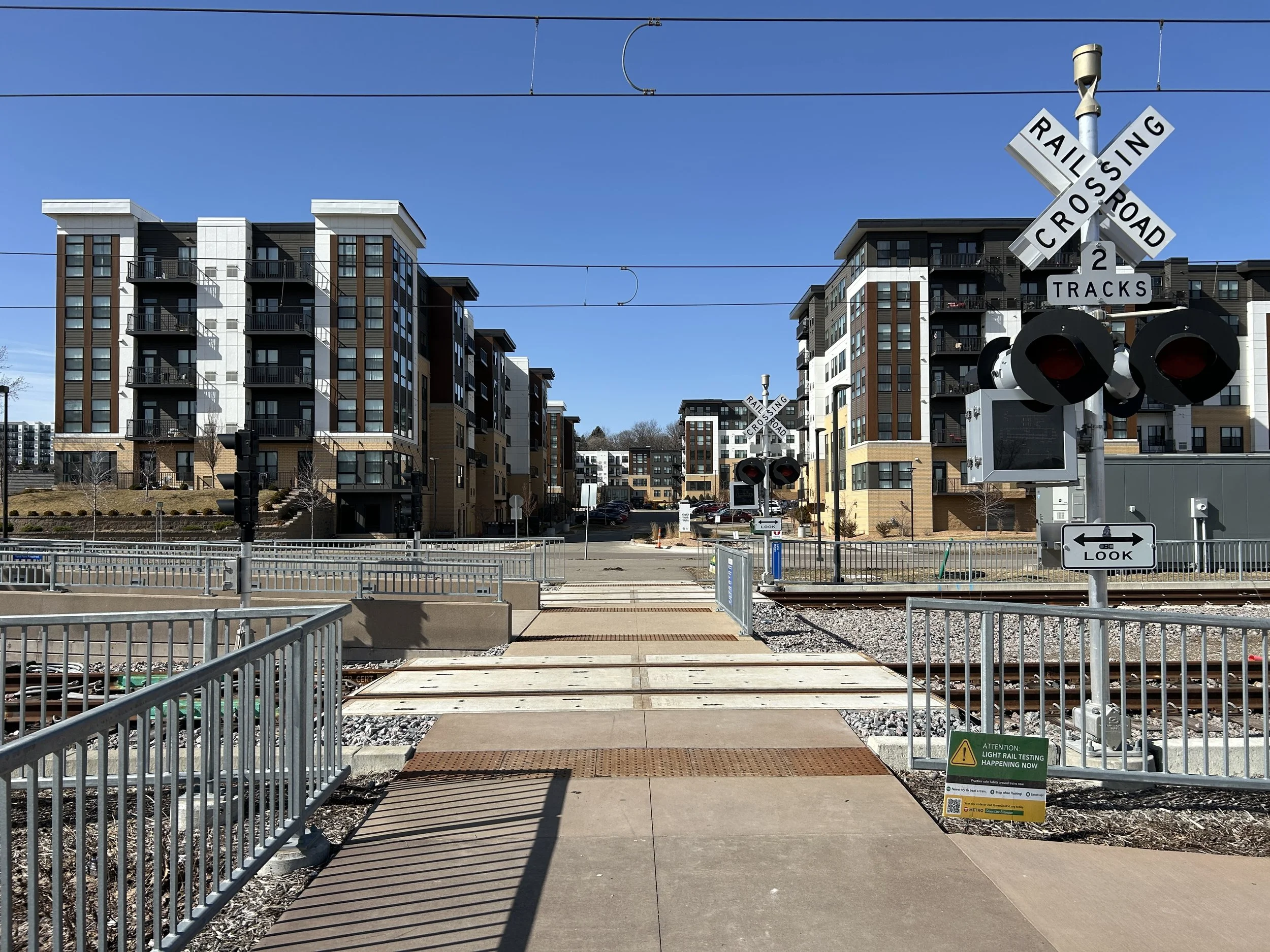 A pedestrian crossing over train tracks with townhouses and apartment buildings in the background, railway crossing signs, and safety signals.