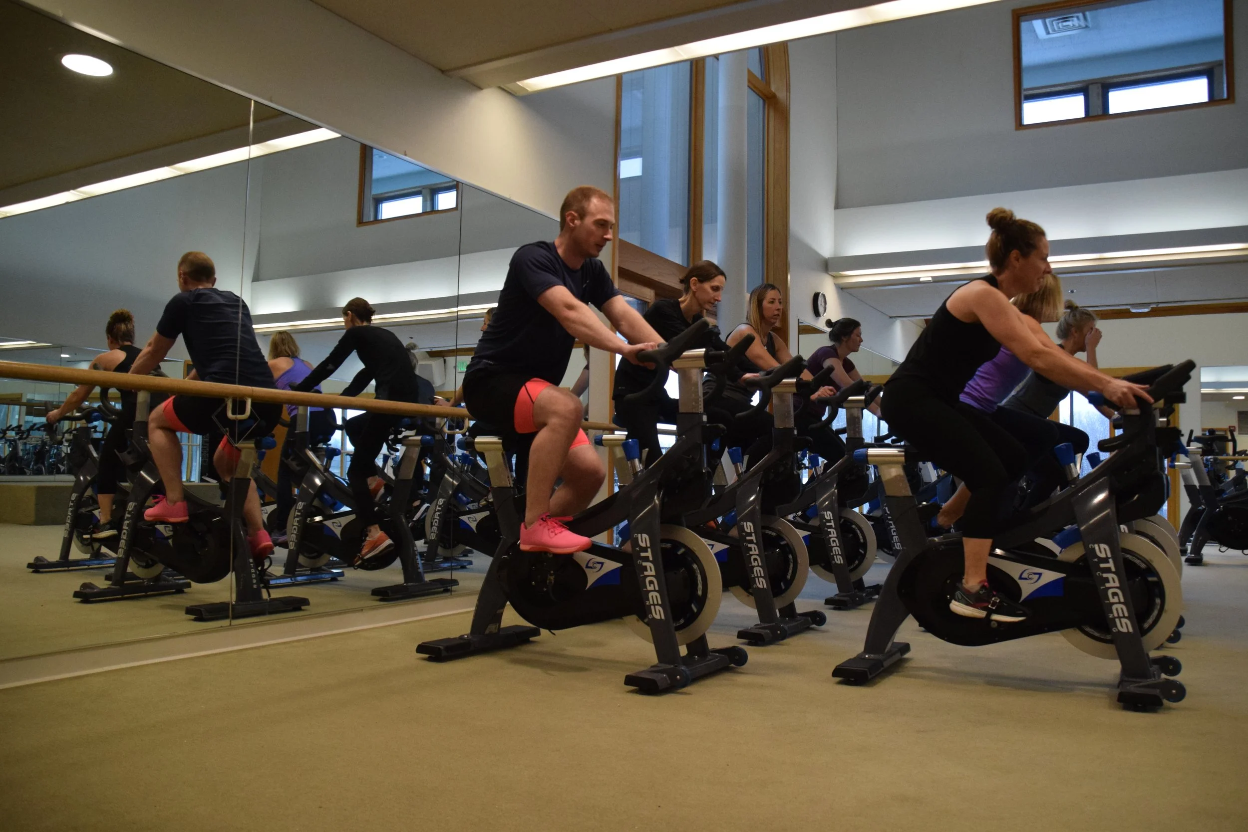 People participating in a spin class on stationary bikes in a gym, with a mirror and window in the background.