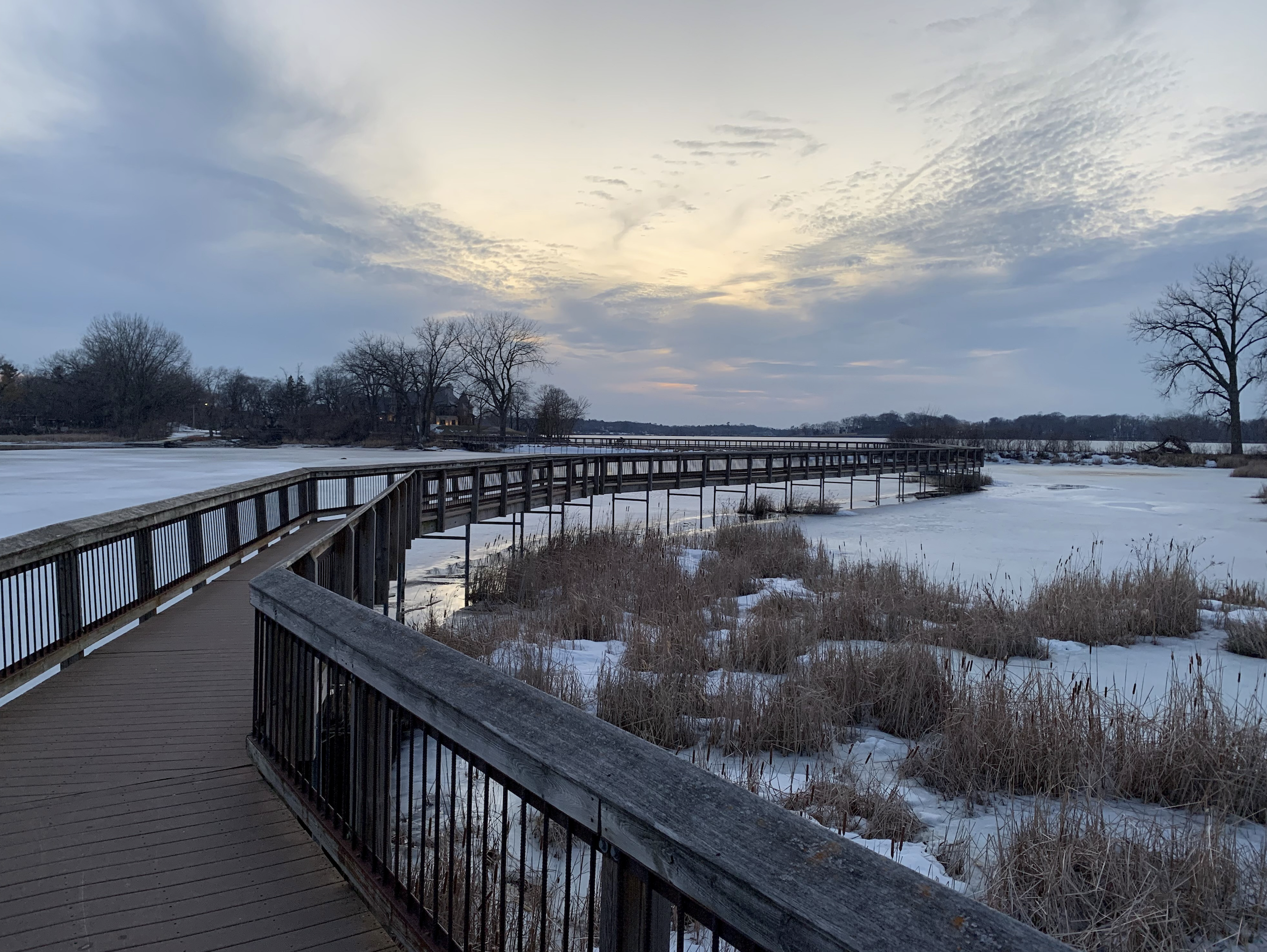 A wooden boardwalk over a partially frozen lake with leafless trees in the background and a cloudy sky at sunset.