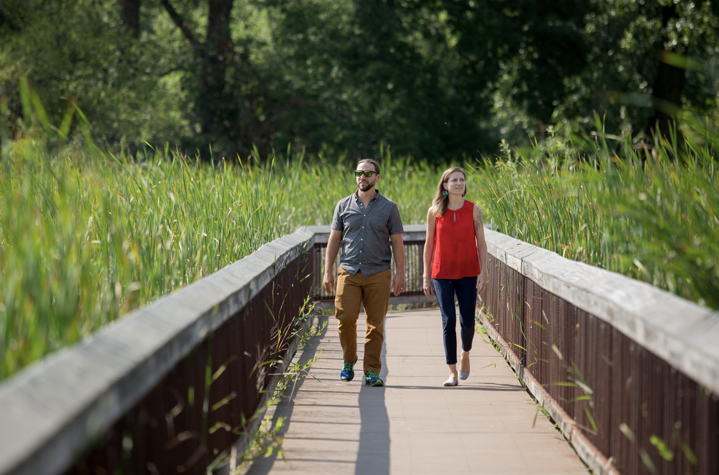 A man and woman walking on a wooden bridge surrounded by tall green grass and trees.