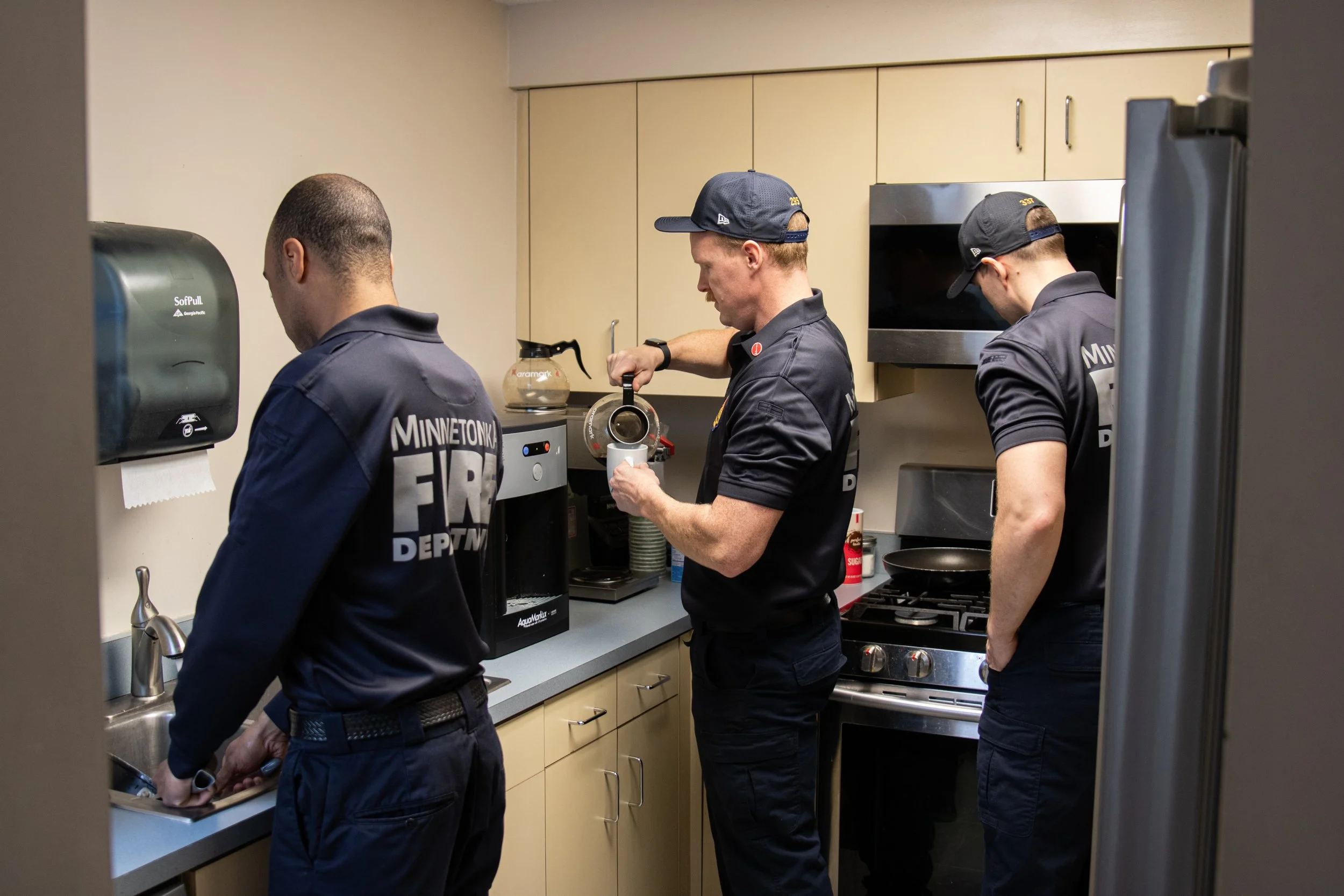 Three uniformed firefighters preparing coffee in a kitchen. One is at the sink, another is pouring water into a coffee maker, and the third is standing with his back to the camera. They are wearing navy blue uniforms with 