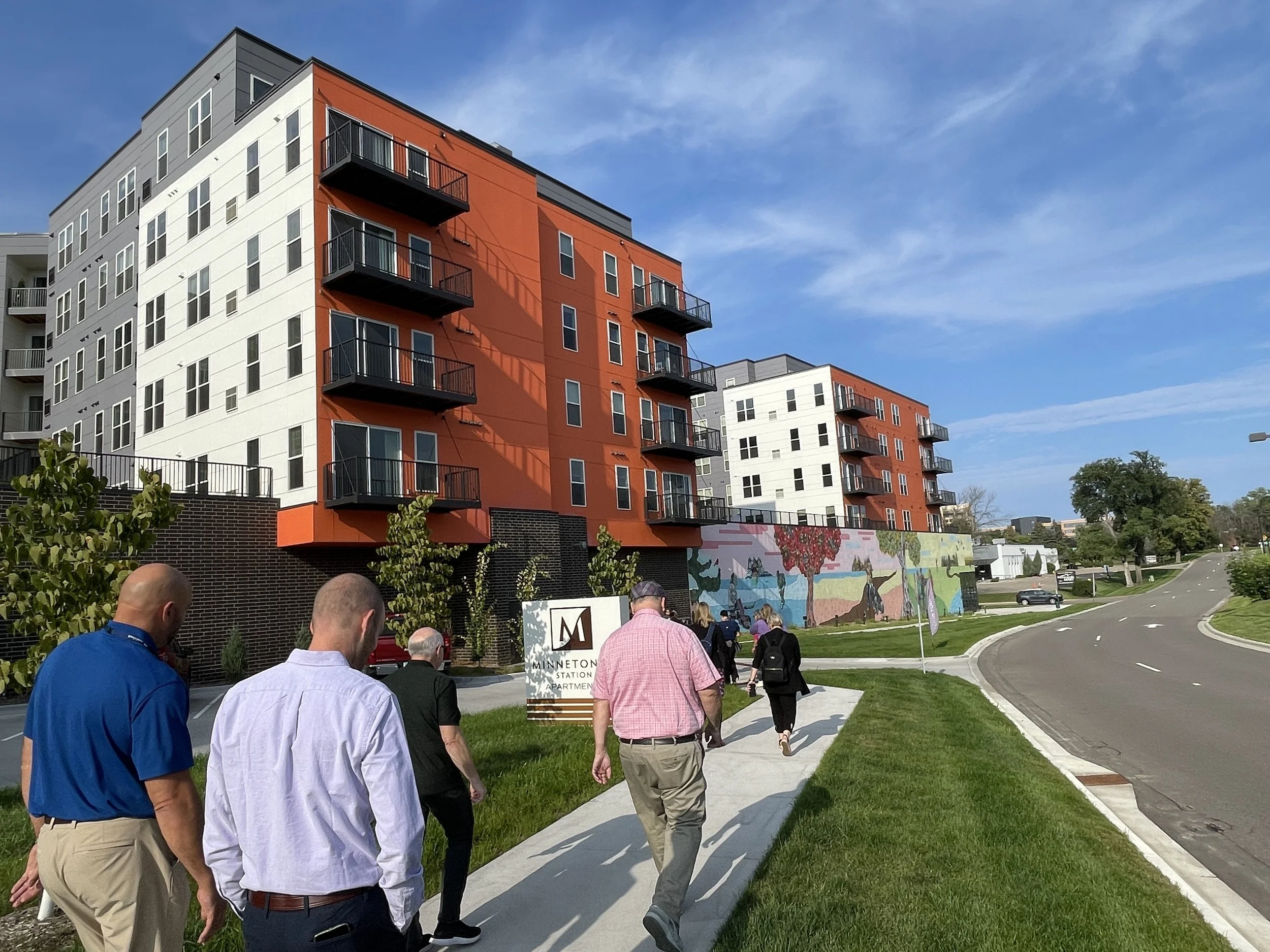 Group of people walking on sidewalk past a colorful modern apartment building with balconies and a mural wall, under a clear blue sky.