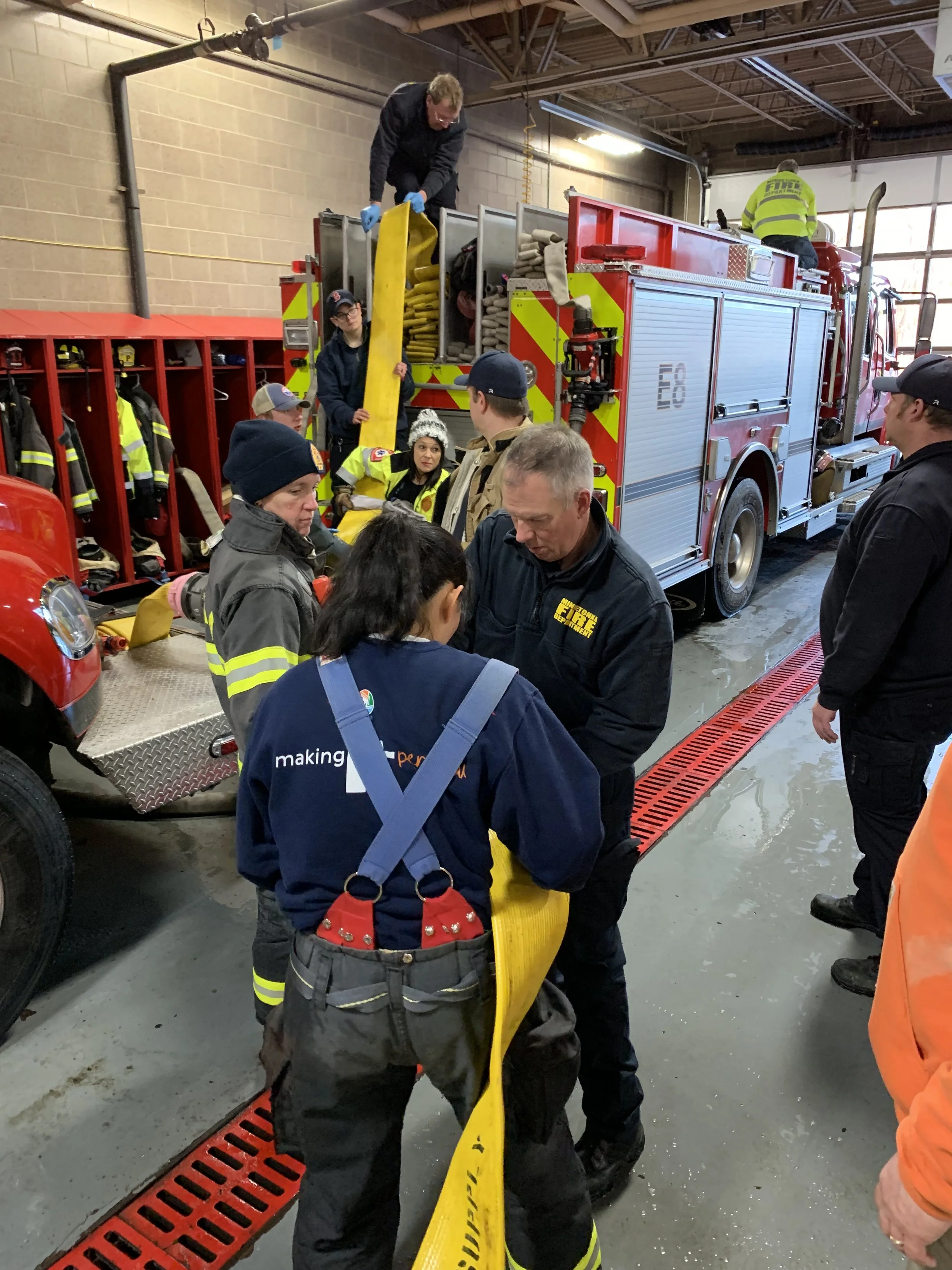 Group of firefighters and trainees inside a fire station, preparing equipment from a fire truck, with some people handling yellow hoses, others observing, and a few working on the truck.