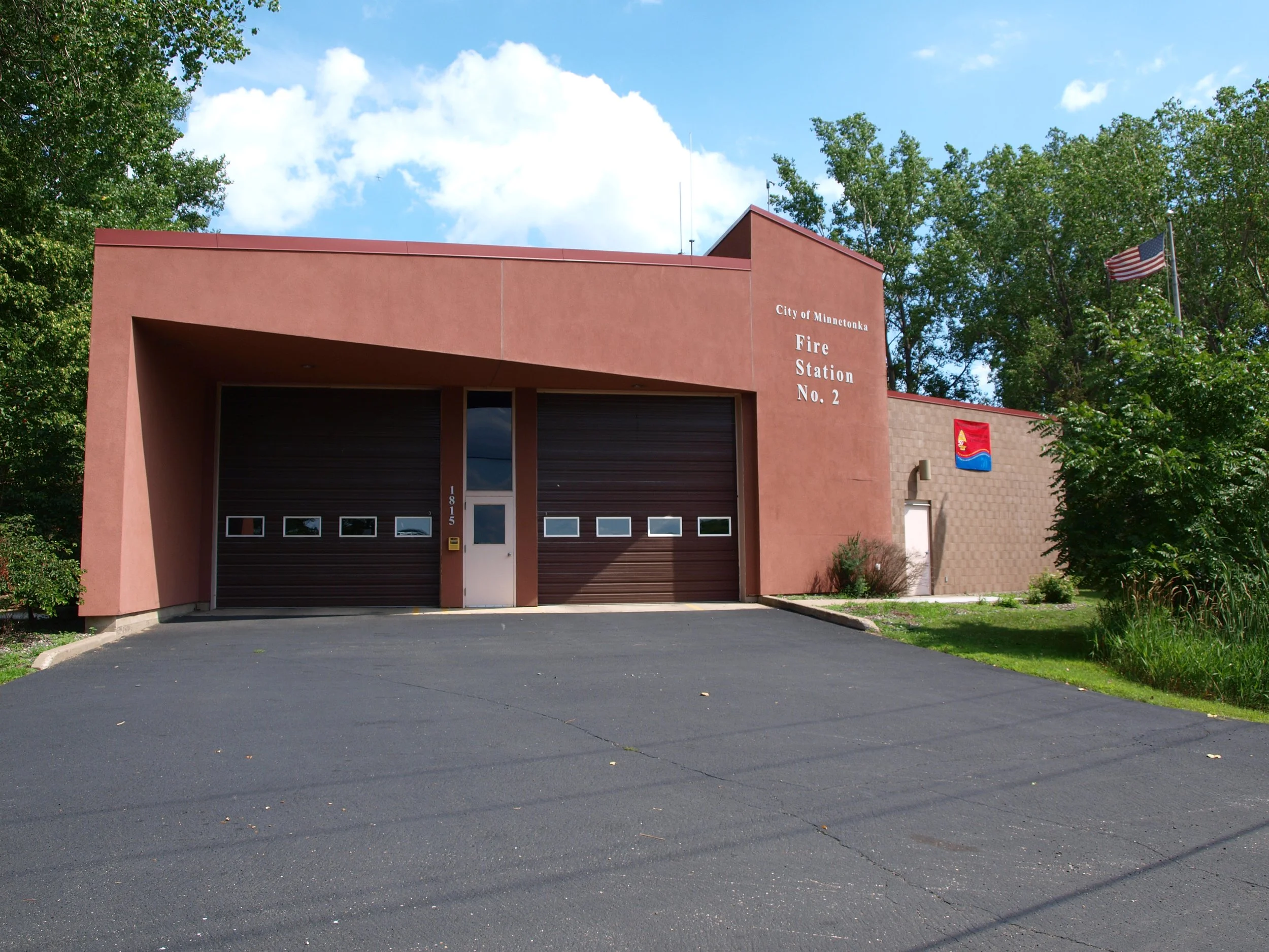 Face of a small fire station building, Minnetonka Fire Station No. 2, with a pinkish-brown facade, two large garage doors, a small white door, and a flagpole with an American flag and another flag nearby. Still and sunny outdoor scene.