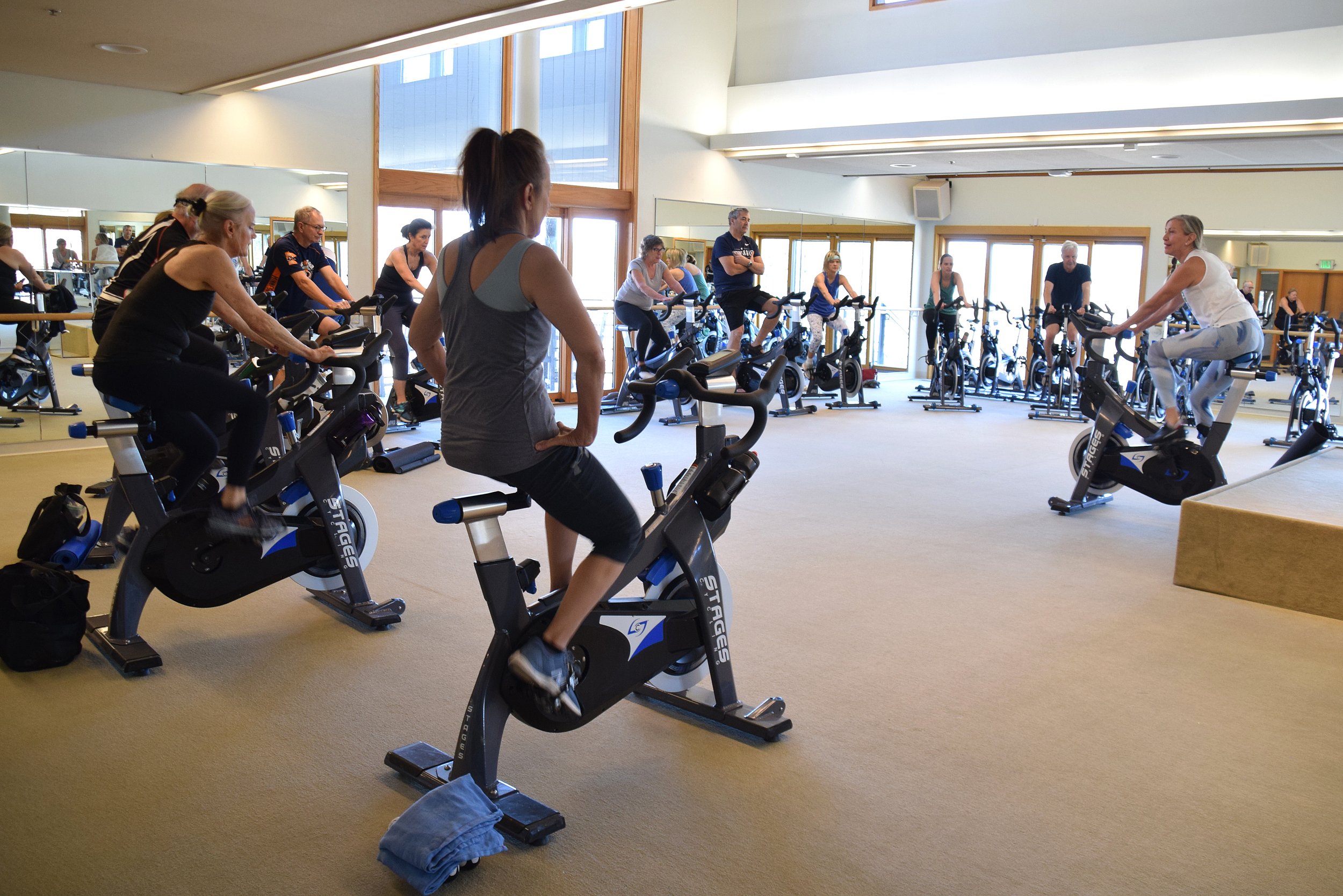 People exercising on stationary bikes in a gym room with large windows and mirrors.