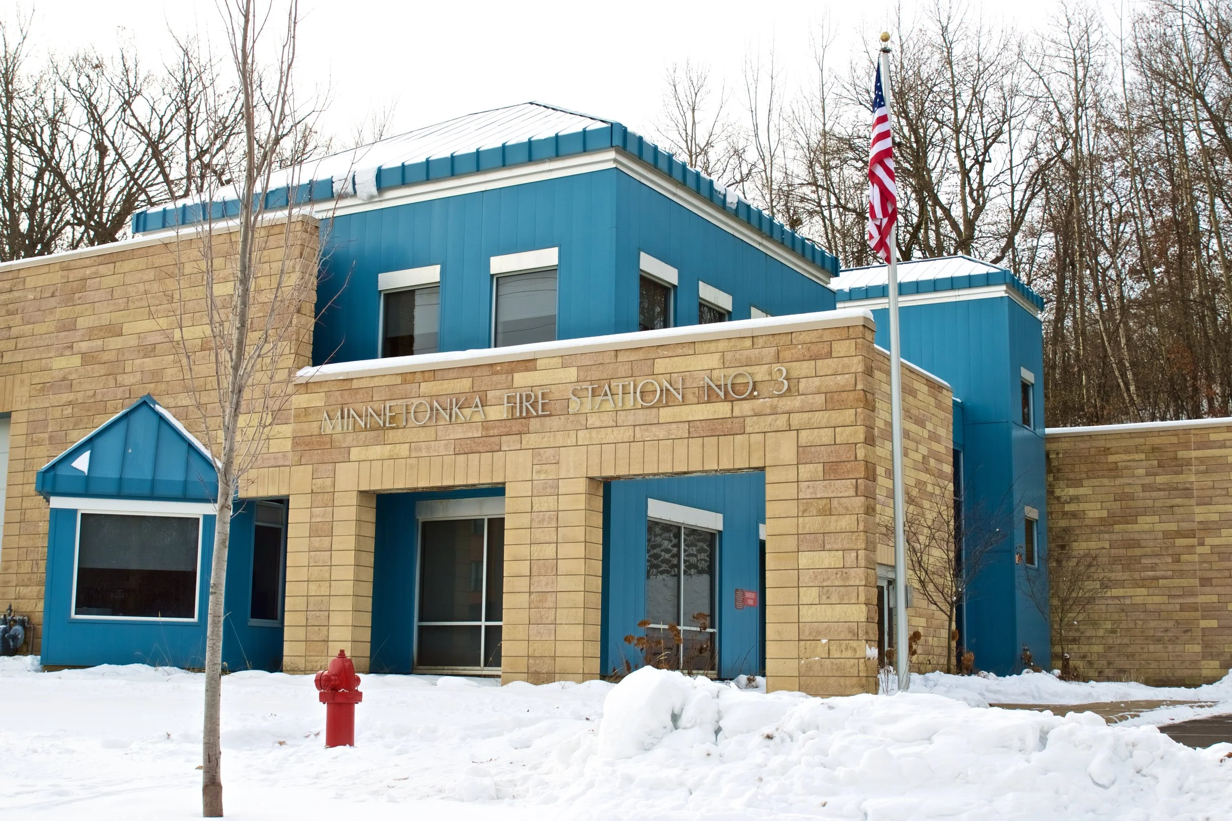 A blue and tan brick fire station building with a sign reading 'Minnetonka Fire Station No. 3', snow on the ground, leafless trees in the background, a red fire hydrant, and an American flag flying on a flagpole.
