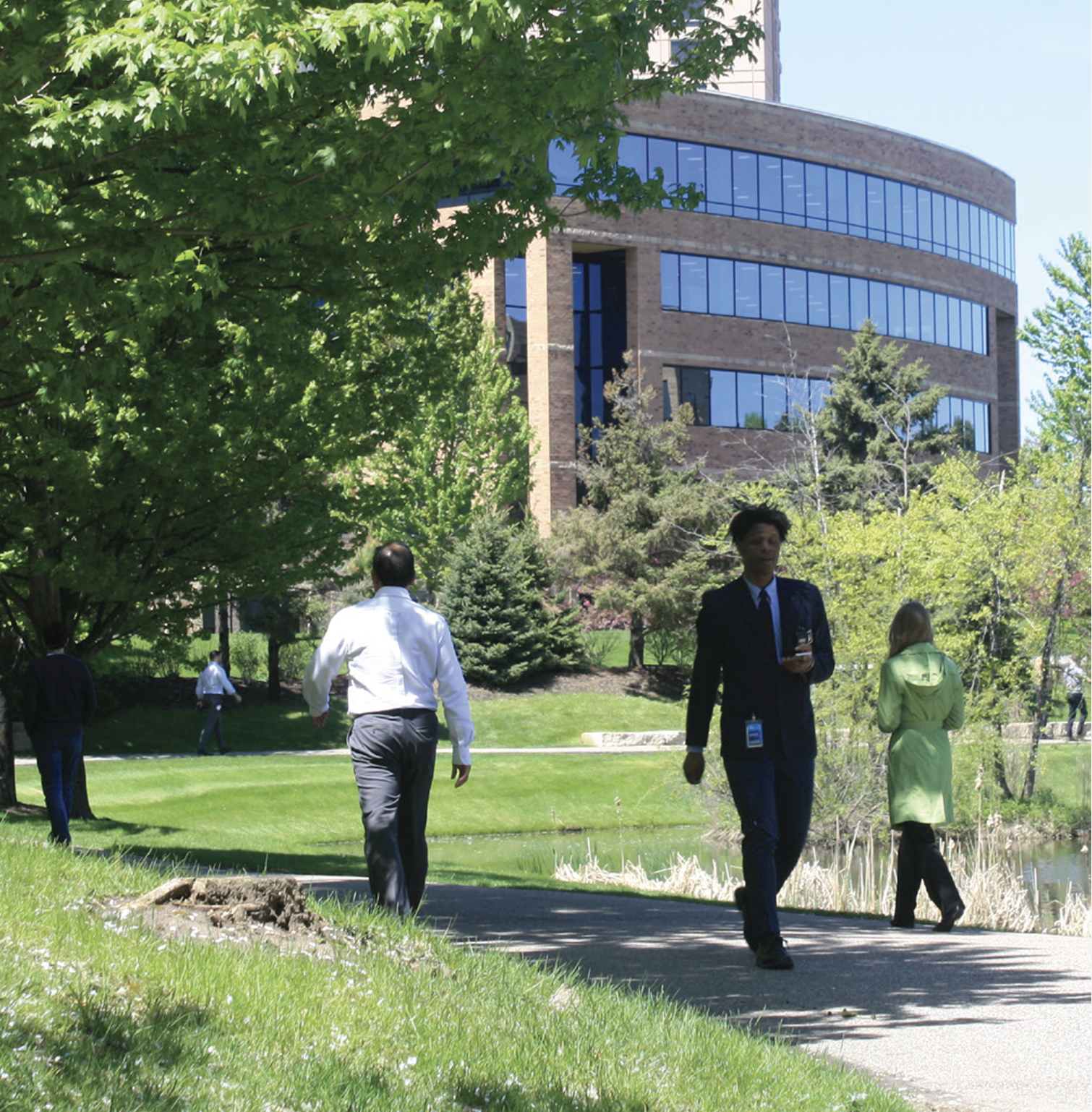 People walking on a path in a park with green trees and grass, and a modern office building in the background on a sunny day.
