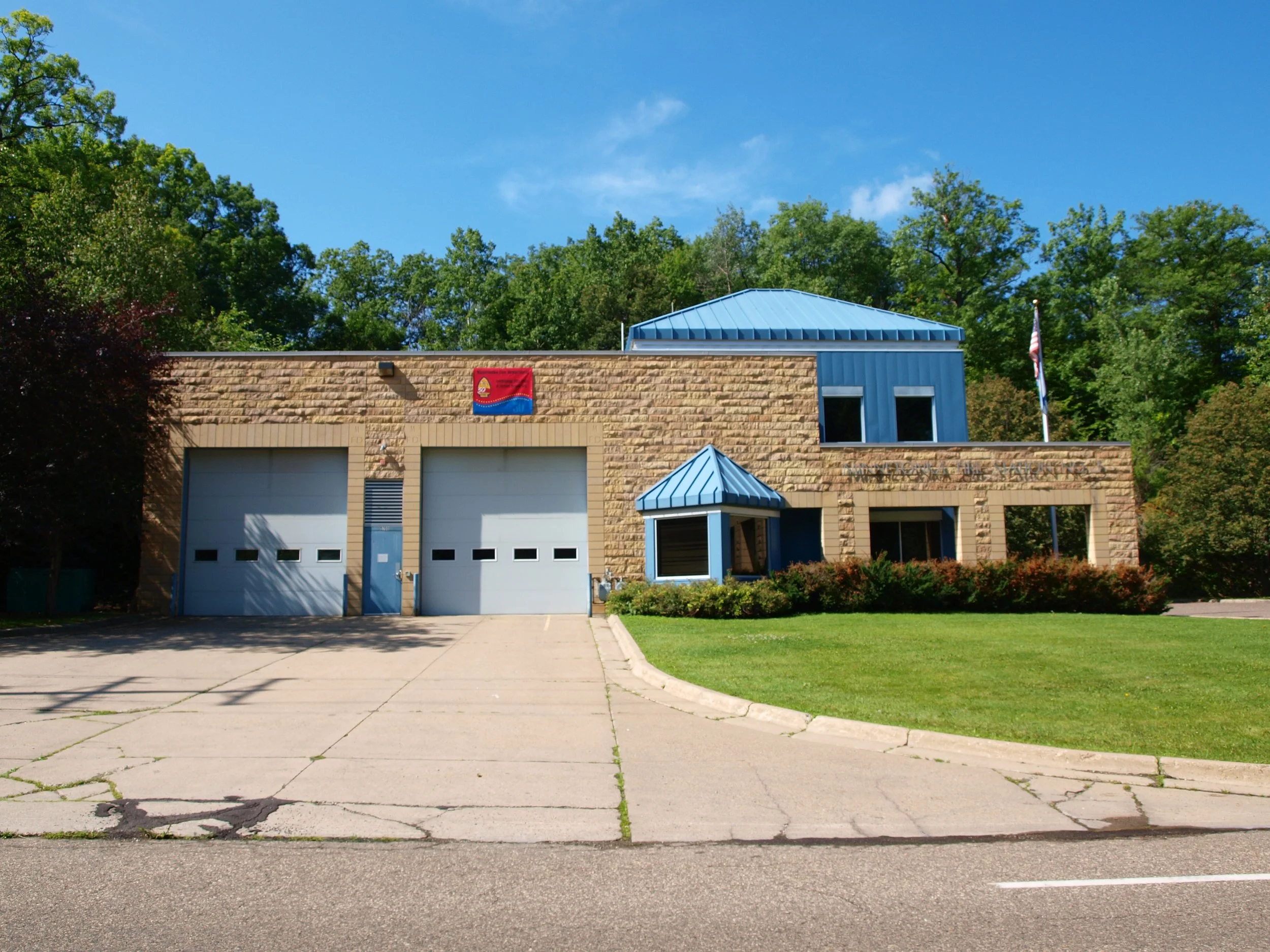 A two-story fire station with a stone facade and blue metal roof, including large garage doors, windows, and a small entrance porch, surrounded by green bushes and trees, with an American flag and a state flag flying in the background under a bright blue sky.