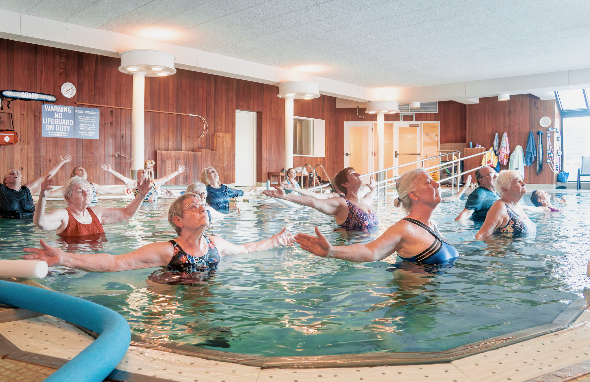 Older adults participating in a water aerobics class in an indoor pool, raised arms in exercise.