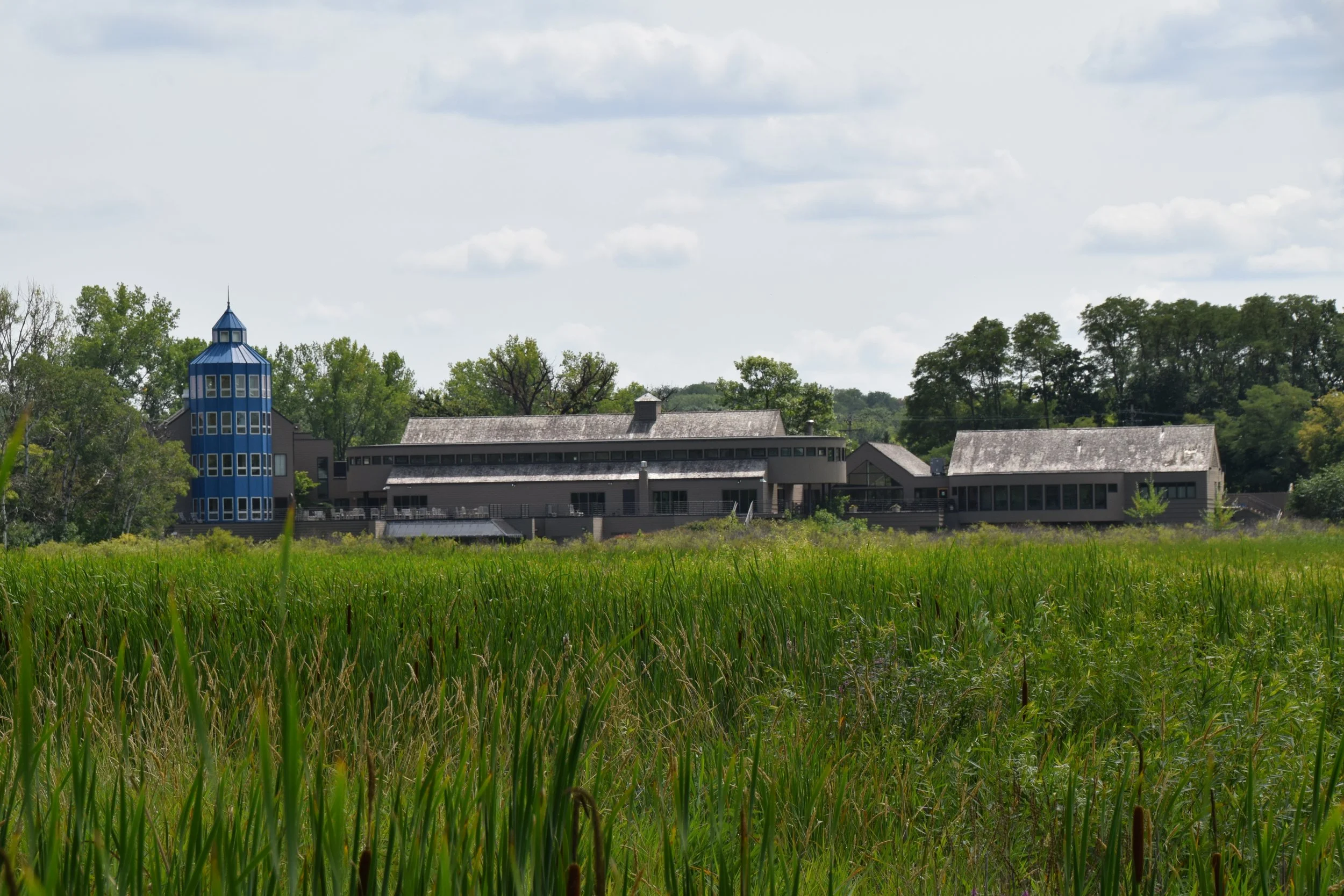 A large building with a blue tower on the left, surrounded by trees and grass, under a partly cloudy sky.