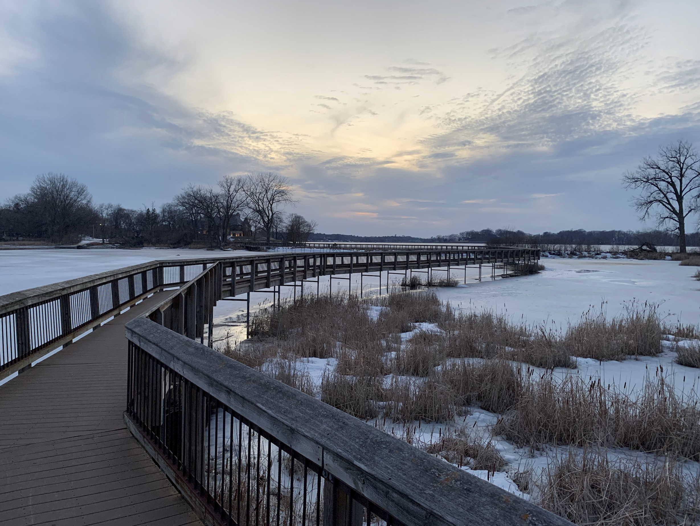 Wooden boardwalk extending over a frozen lake with dry grass and leafless trees in winter under a cloudy sky at sunset.