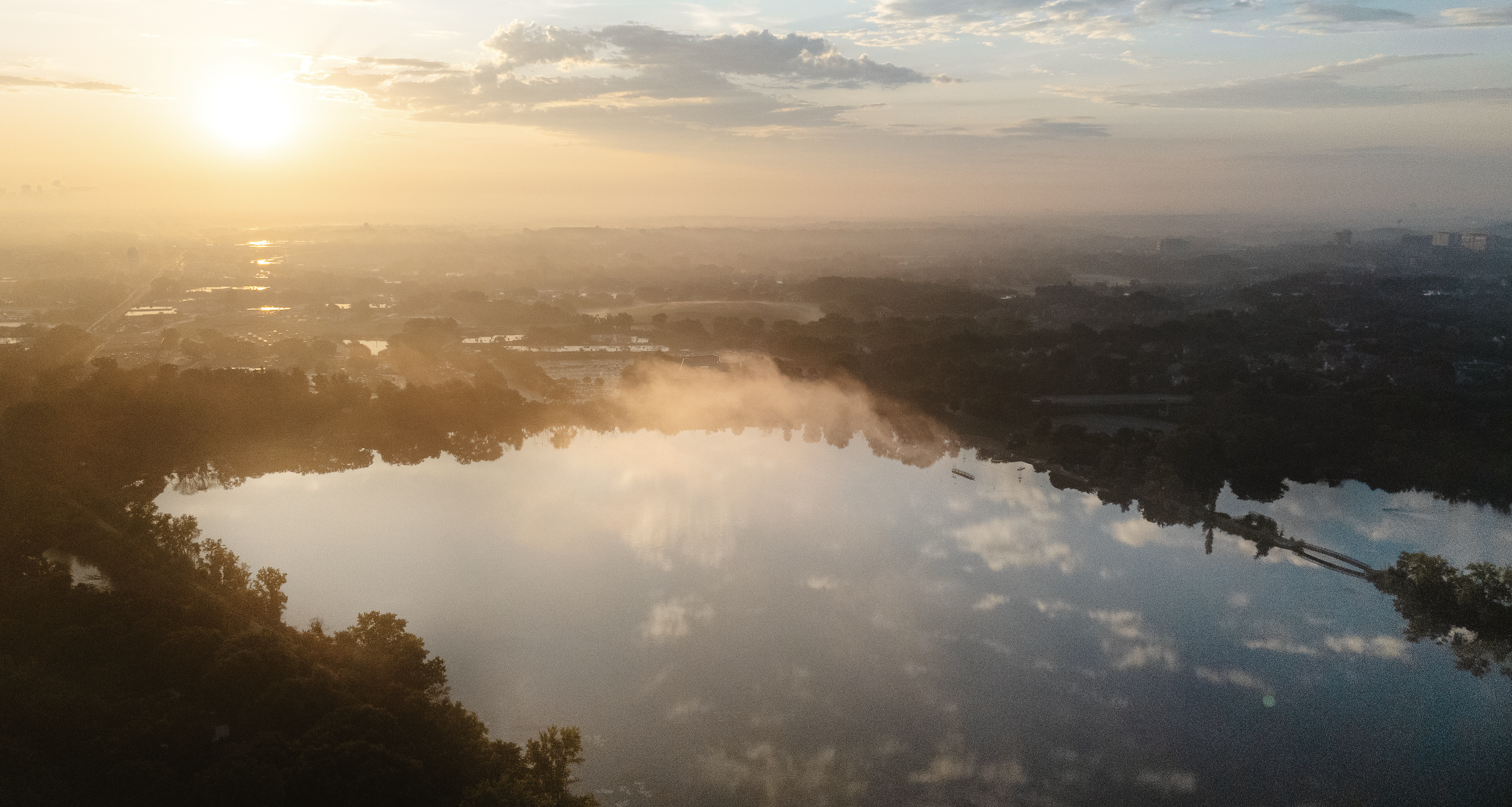 Aerial view of a lakeside landscape at sunrise with the sun shining over the water, reflecting clouds and sky, surrounded by trees and distant city buildings.