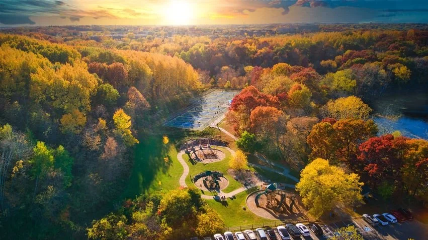 Aerial view of a park during sunset with trees in fall colors, walking paths, a tennis court, a playground, and a parking lot.