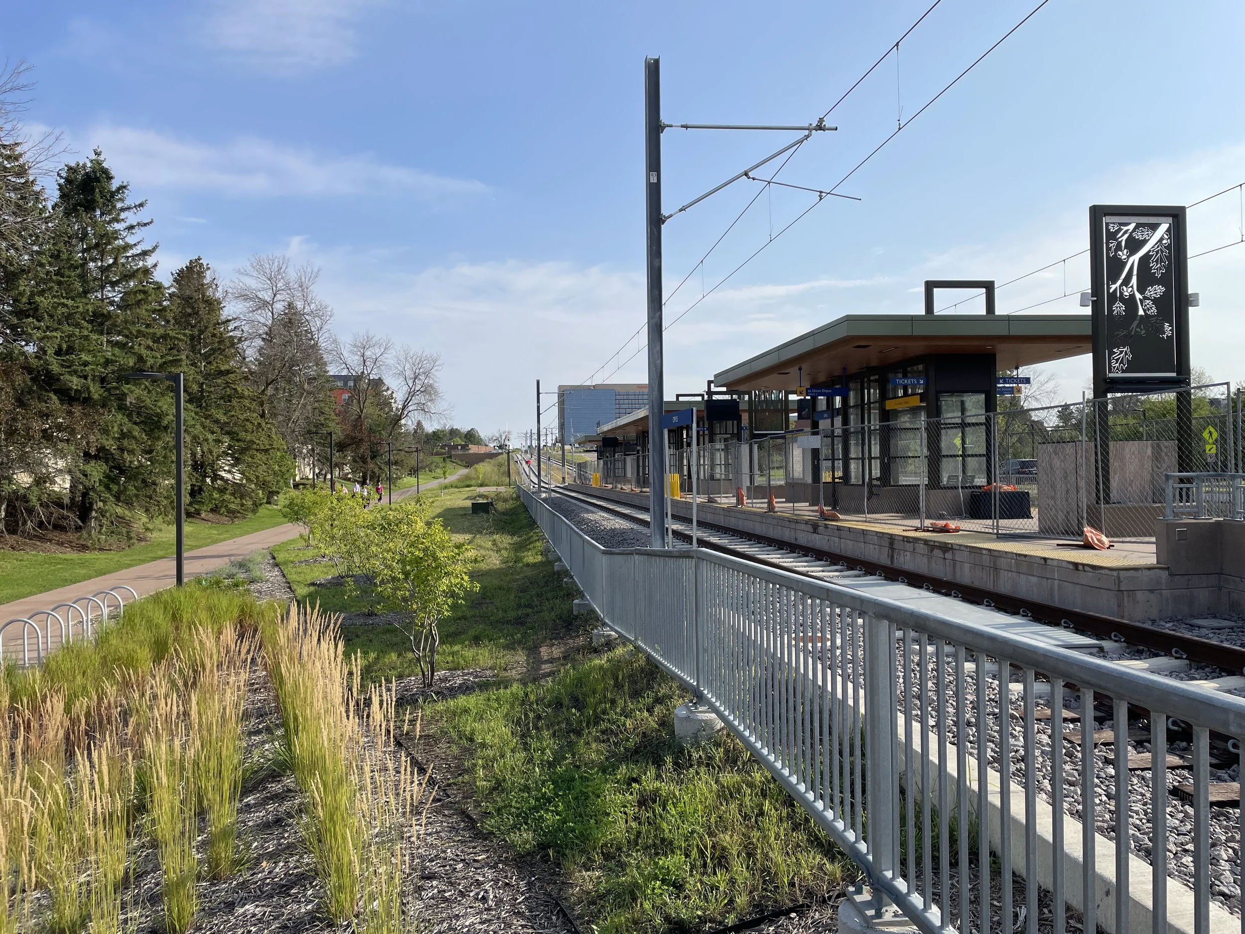 An empty train station platform with a shelter and ticket machines, surrounded by a fence, next to a park with trees and a walking path, under a blue sky with some clouds.