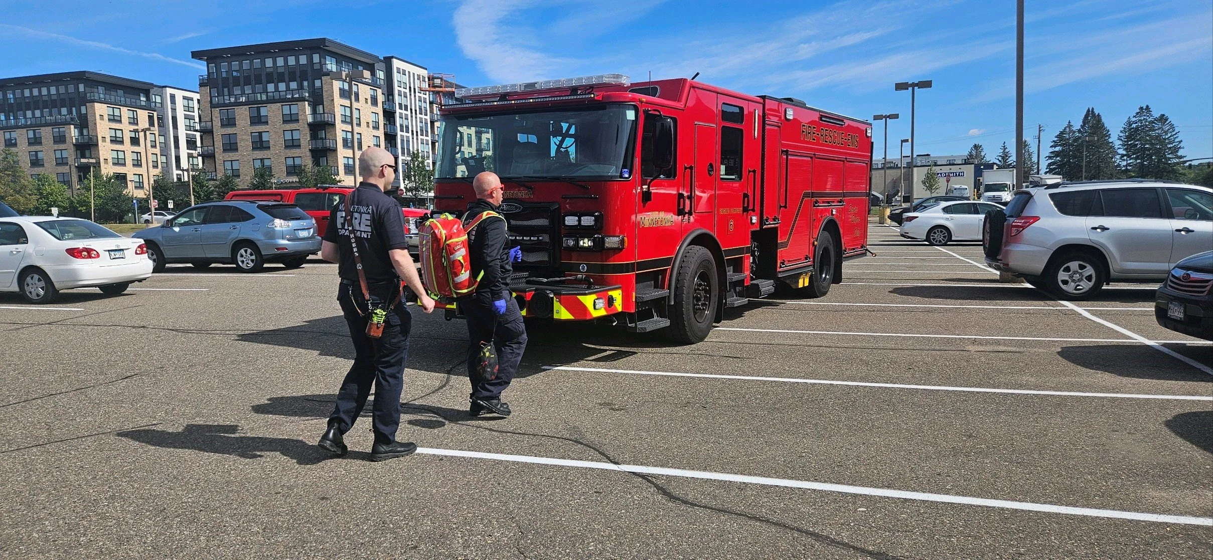 Two firefighters in black uniforms with equipment walking near a red fire truck in a parking lot with parked cars and apartment buildings in the background.