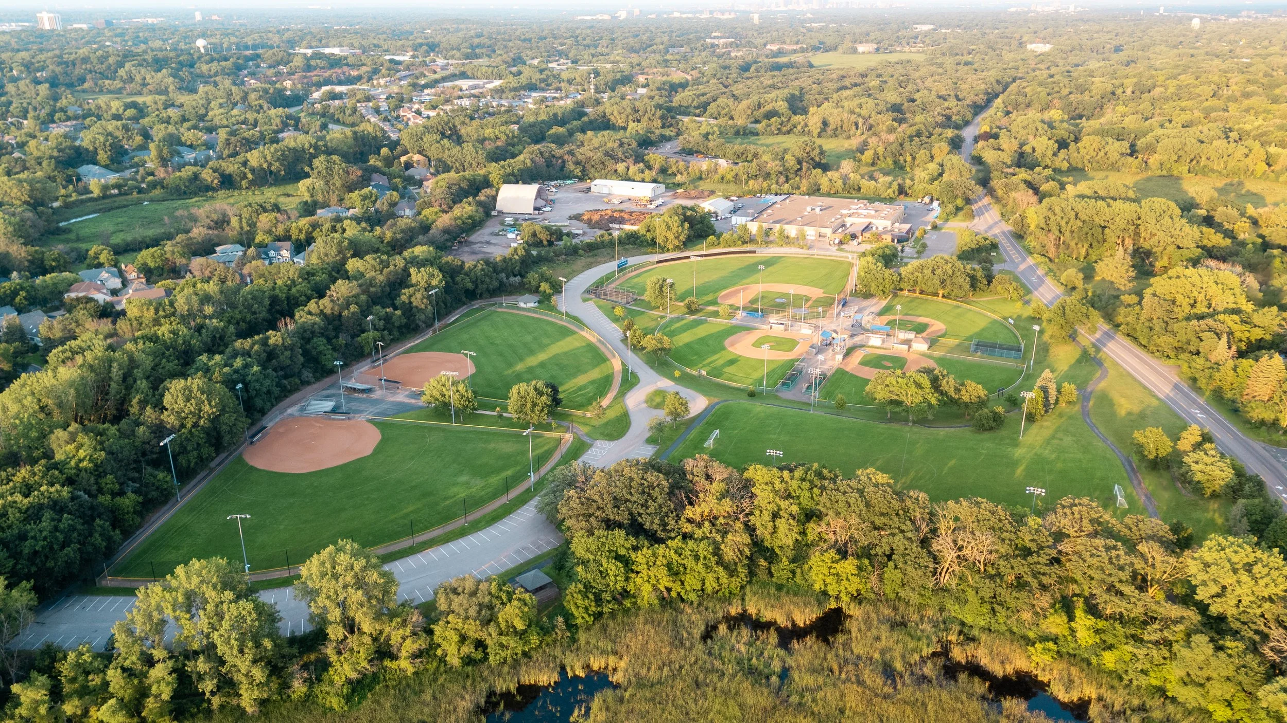 Aerial view of a baseball and softball complex with multiple fields, parking lots, surrounded by green trees and residential neighborhoods.