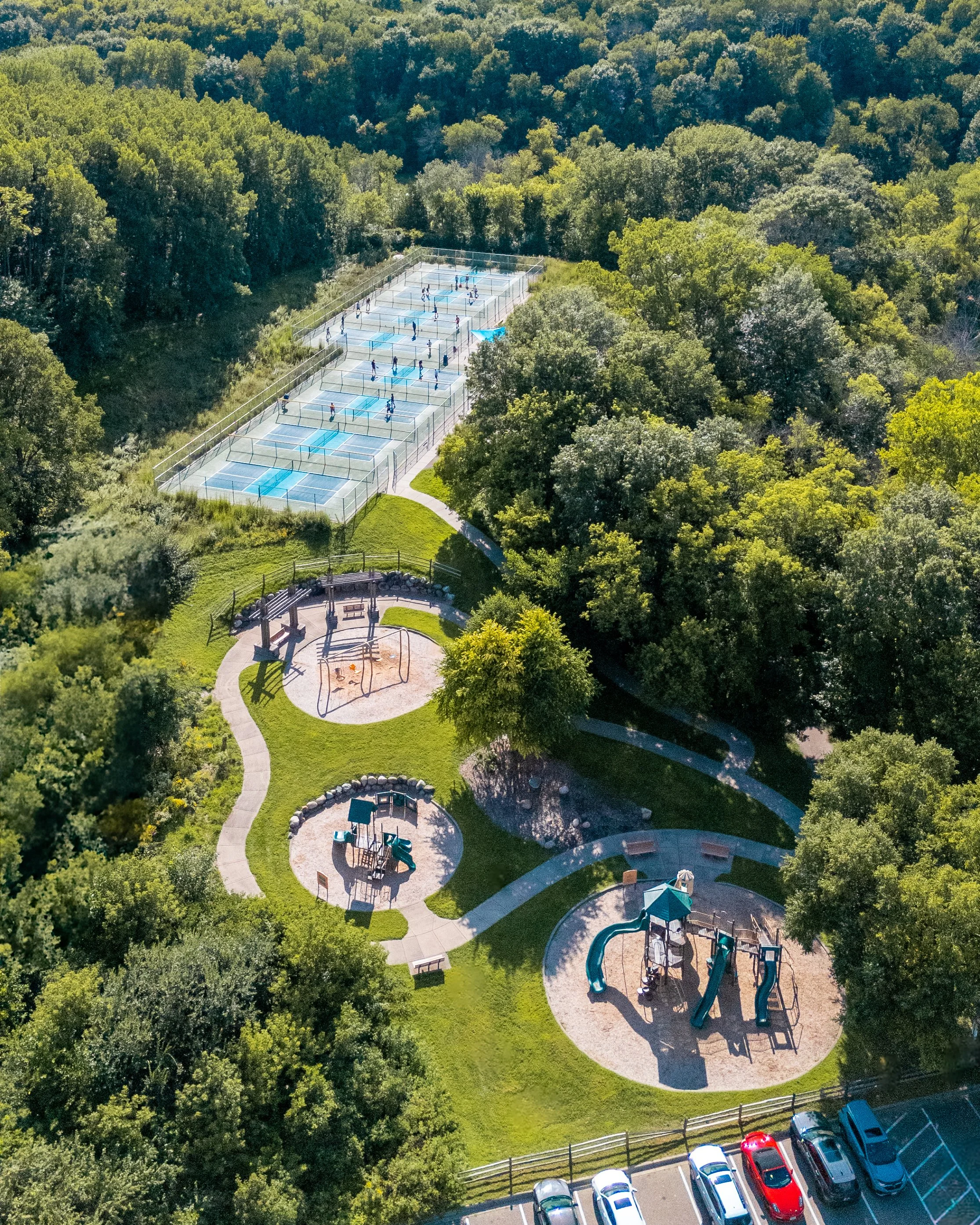 Aerial view of a park with several playgrounds and tennis courts surrounded by dense trees, with parked cars at the bottom.