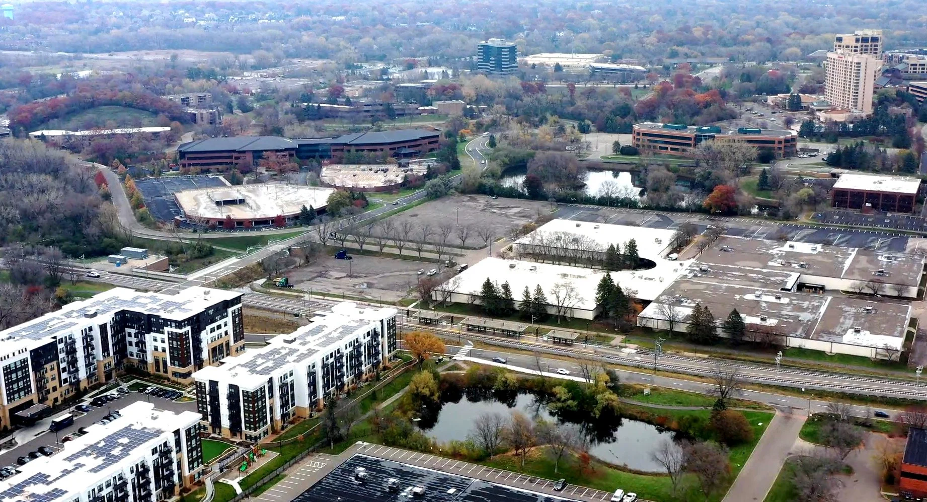 Aerial view of an urban area at Opus with residential buildings, parking lots, a small pond, and many trees, some with autumn foliage.