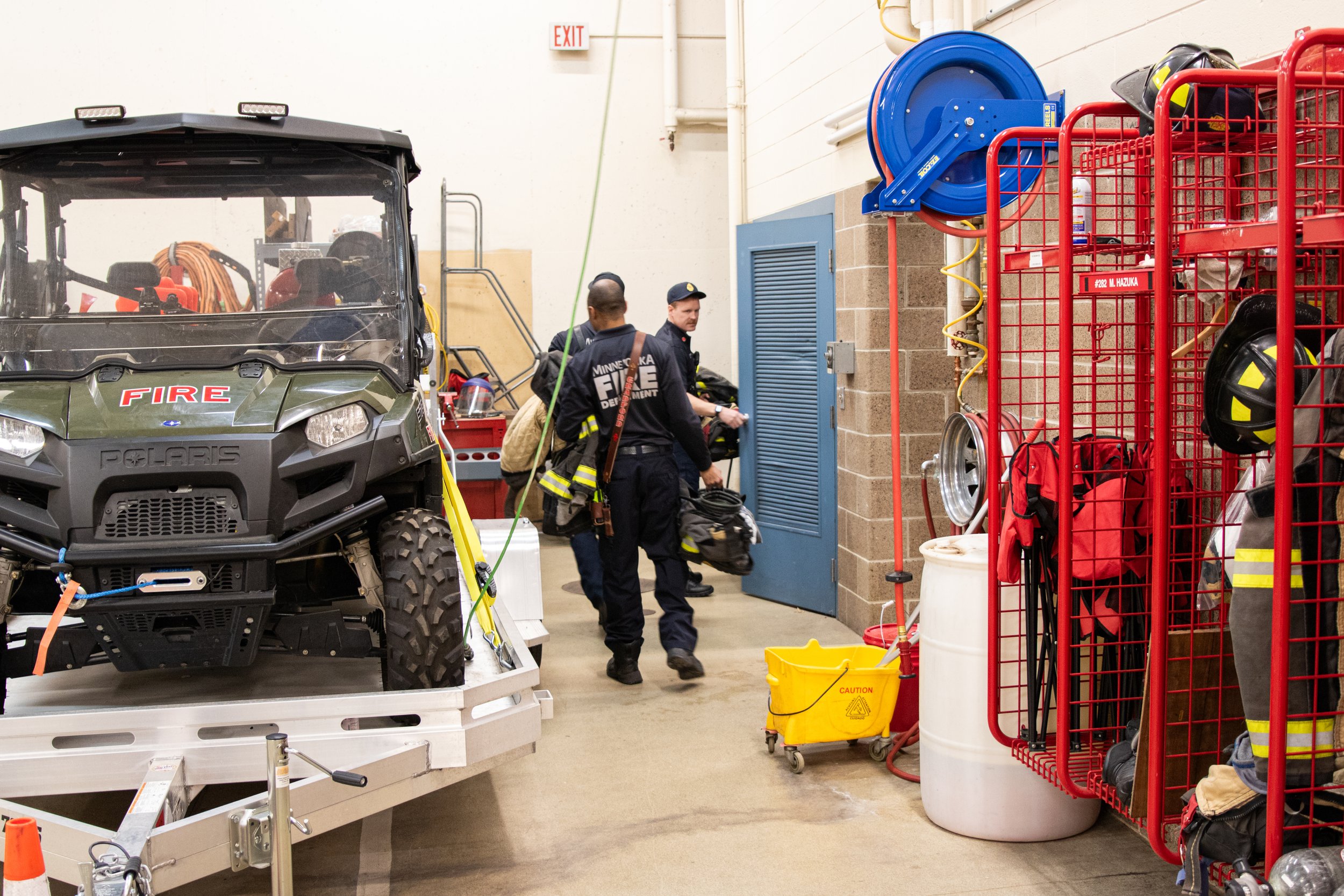 Firefighters in a fire department garage, with equipment including helmets, backpacks, and a Polaris utility vehicle, and firefighting gear stored on red carts.