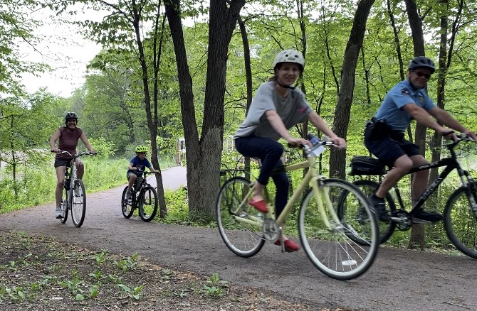 Four people riding bikes on a tree-lined forest trail, wearing helmets and casual clothing.