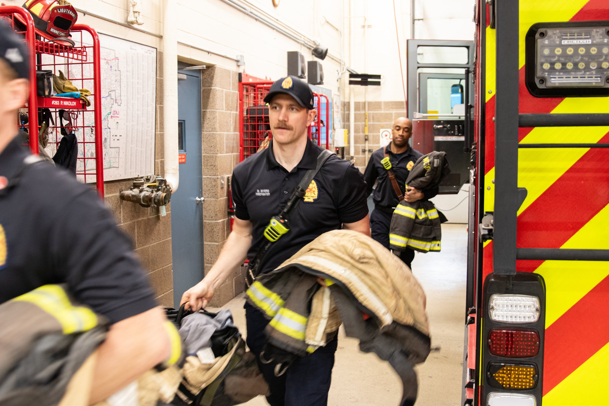 Firefighters inside a fire station, preparing equipment, with a firefighter partially visible in the foreground, a firefighter holding a bag, and another firefighter in the background near a fire truck.