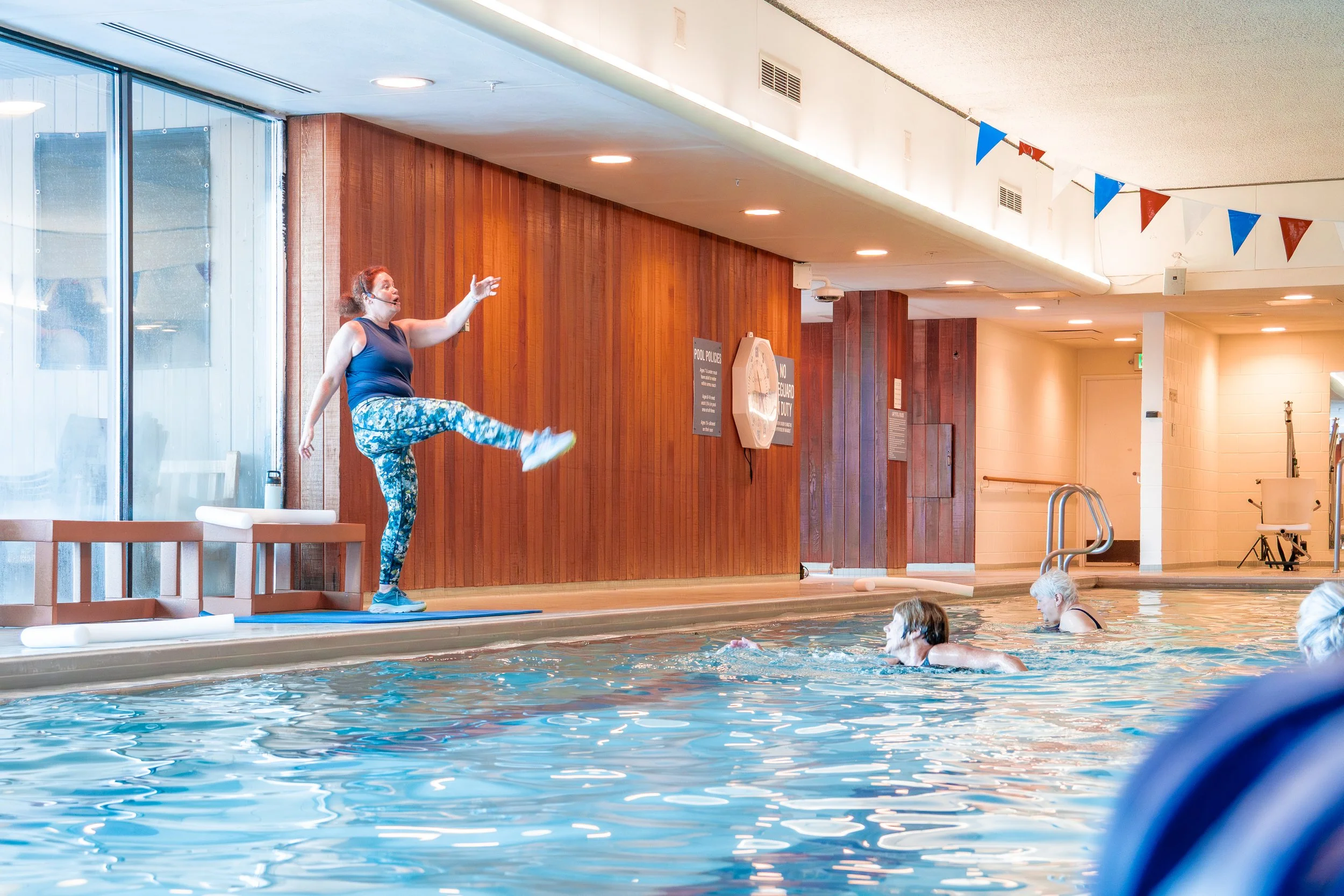 An instructor leading a water aerobics class in an indoor swimming pool, demonstrating a high-knee exercise on the pool deck while participants follow her in the water.