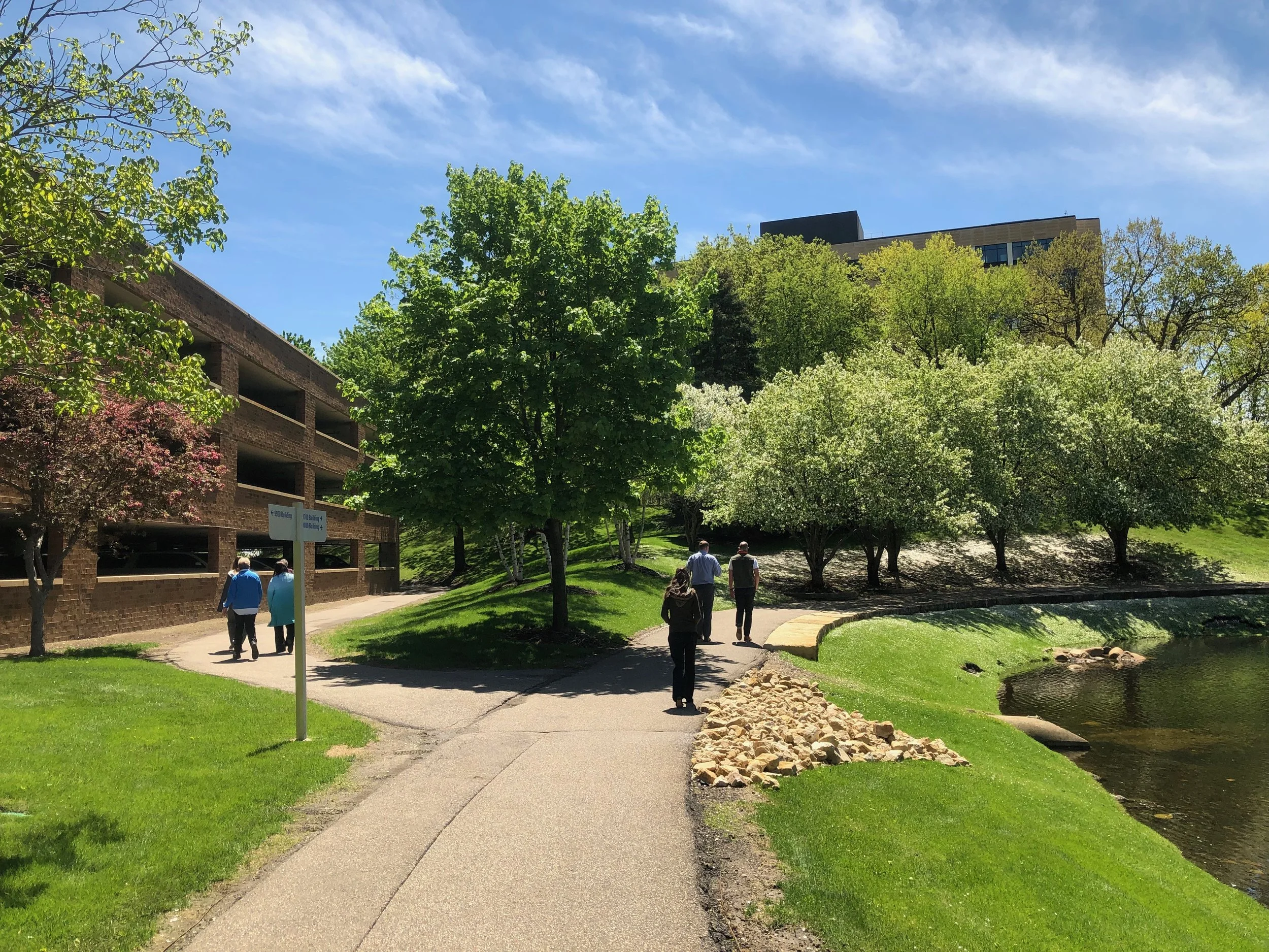 A group of five people walking on a paved path in a park with green grass, trees, and a pond. A brick parking garage is on the left, and a modern building is visible in the background on a sunny day with a bright blue sky.