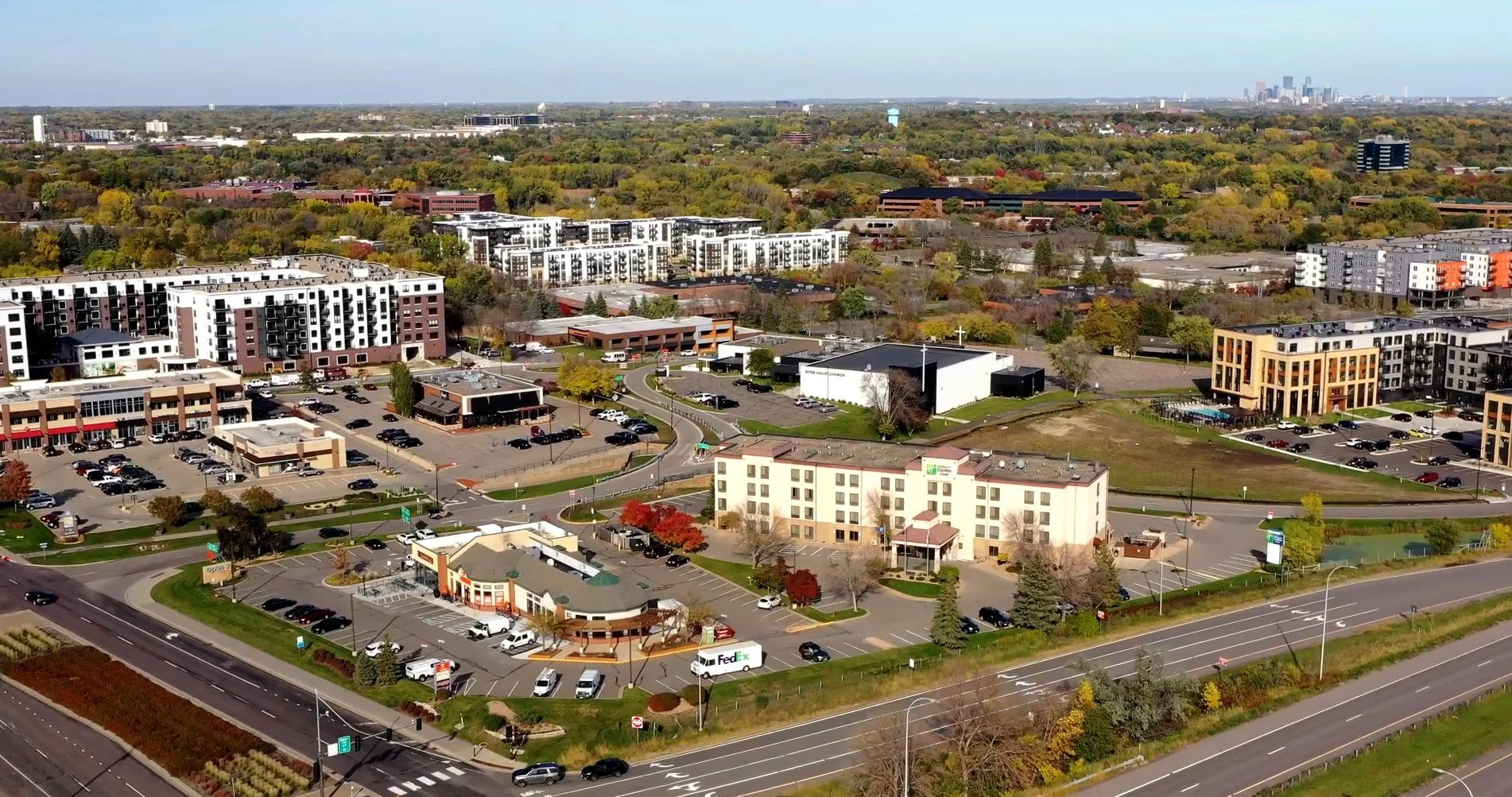 Aerial view of a commercial and residential area at Opus with a mix of buildings, parking lots, trees, and a highway in the foreground, with a city skyline in the distance.