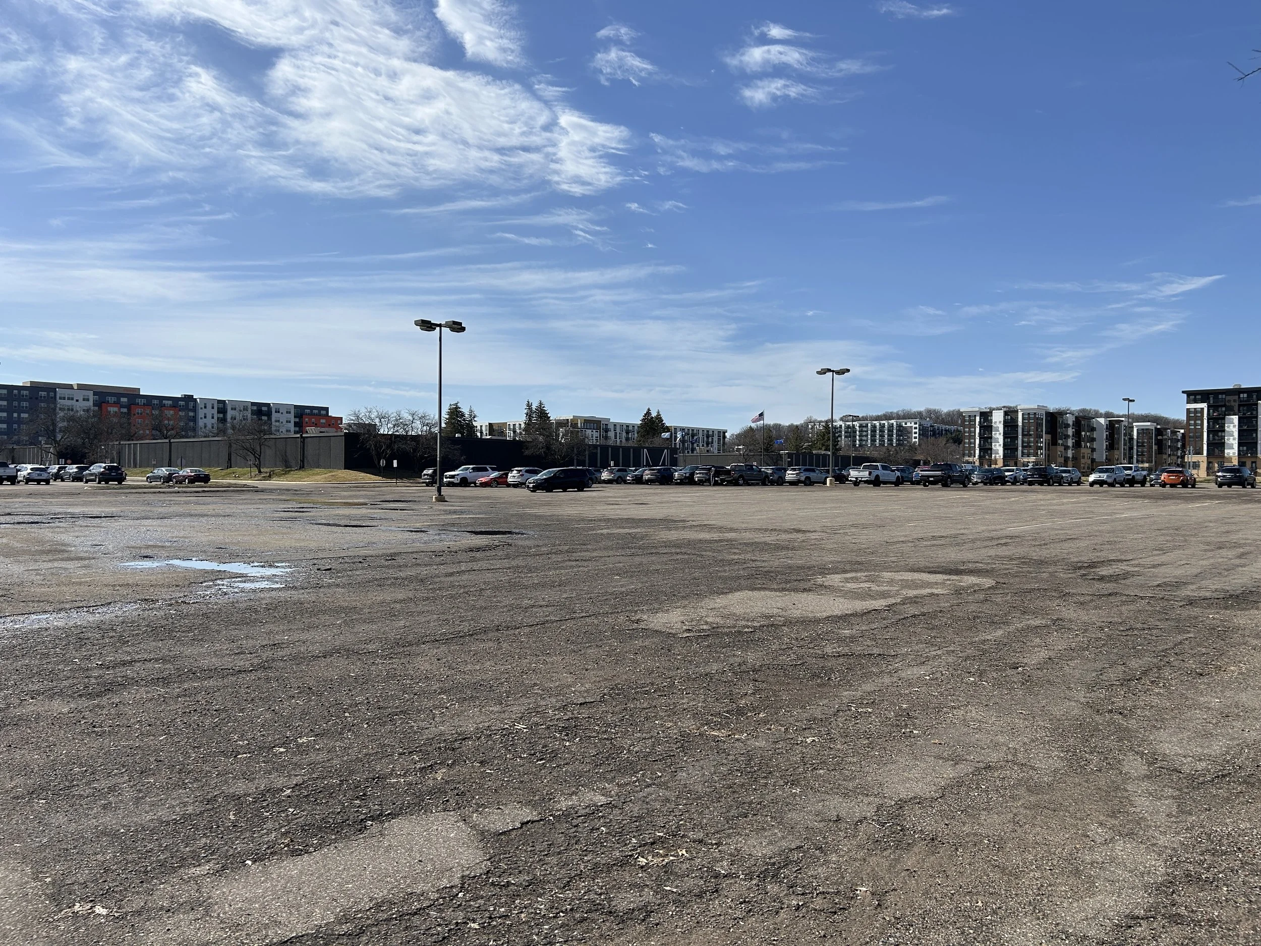 Wide shot of an empty parking lot with a cityscape of modern apartment buildings in the background and a bright blue sky with scattered clouds above.