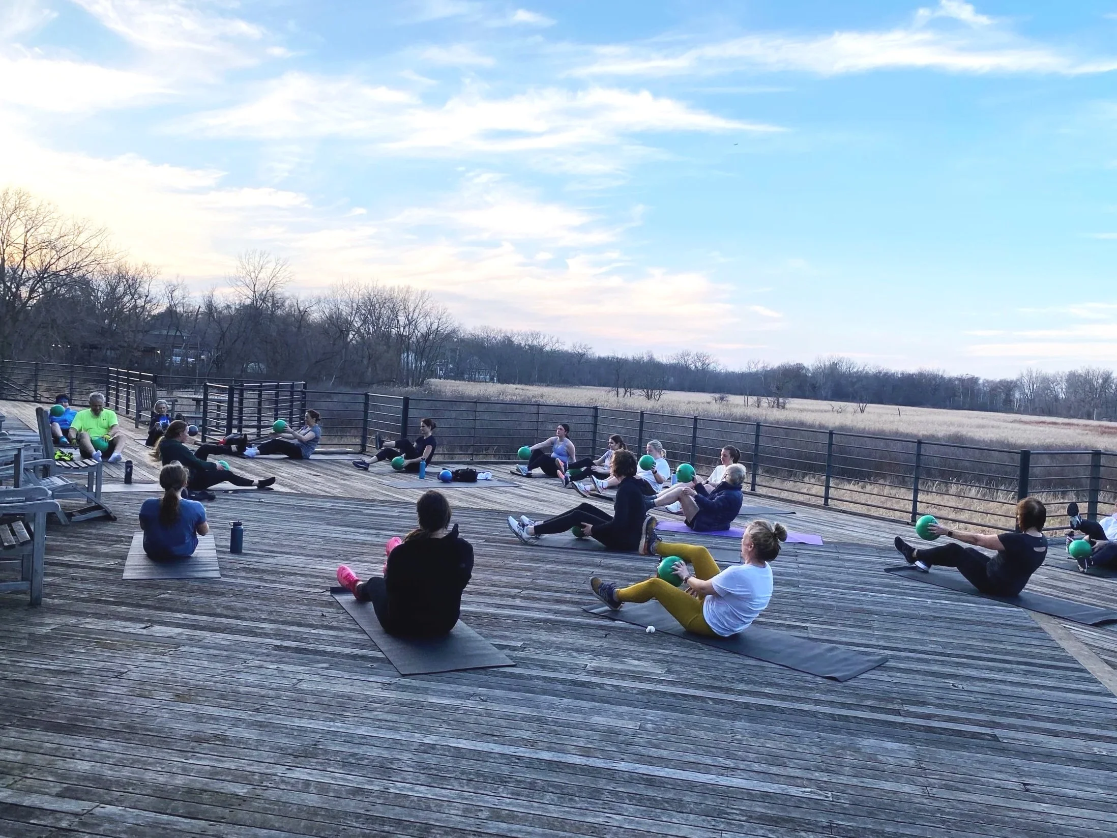 People participating in an outdoor fitness class on a wooden deck, sitting on mats with green exercise balls, under a partly cloudy sky.