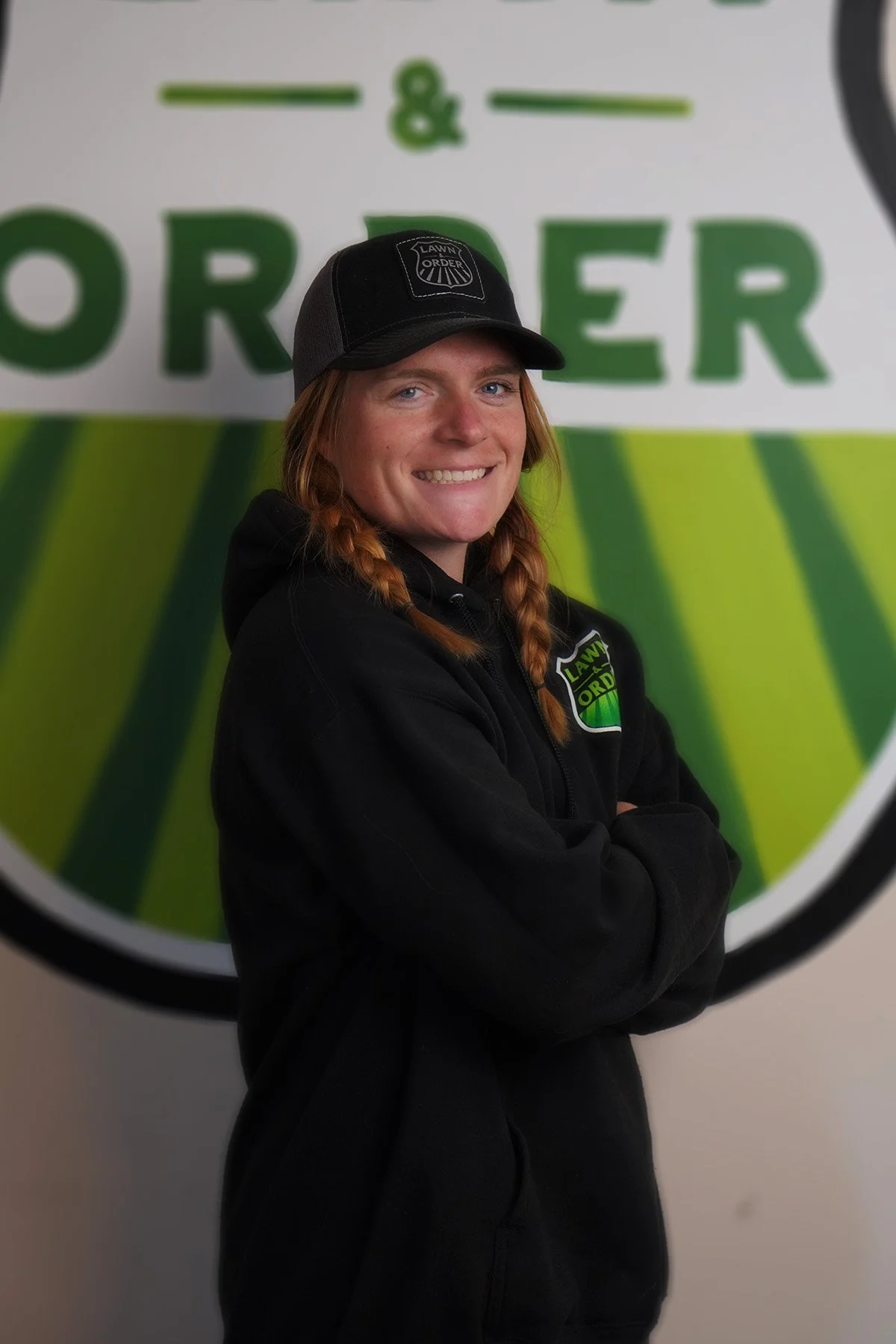 A woman with red hair in braids, wearing a black hoodie and a black cap, standing with arms crossed in front of a large sign that says 'Lawn & Order'.
