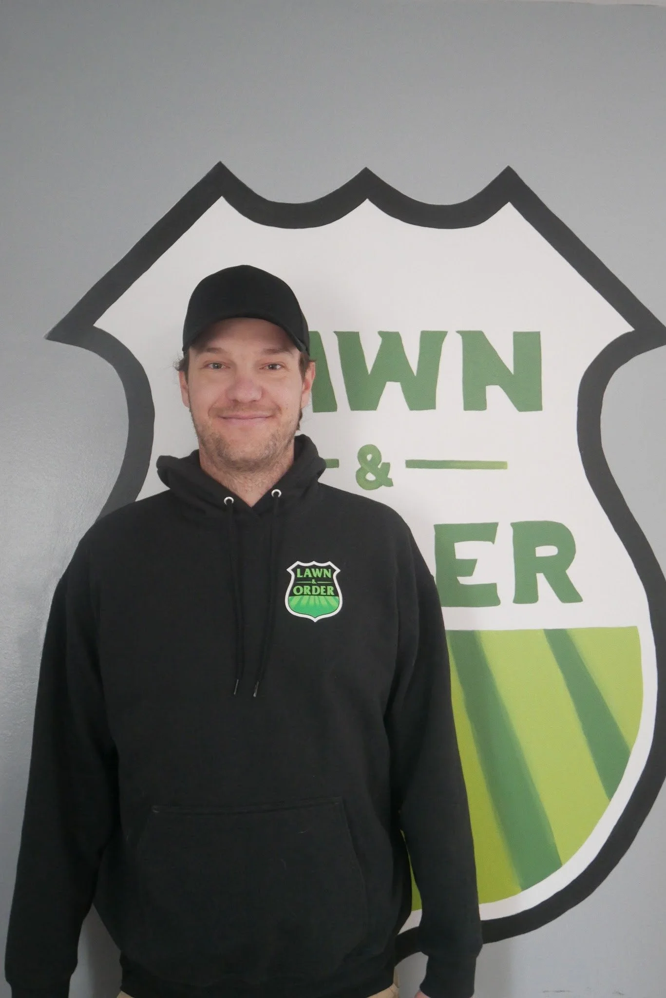 A man wearing a black hoodie and cap standing in front of a wall with a logo that says "Lawn & Order" in green and black.