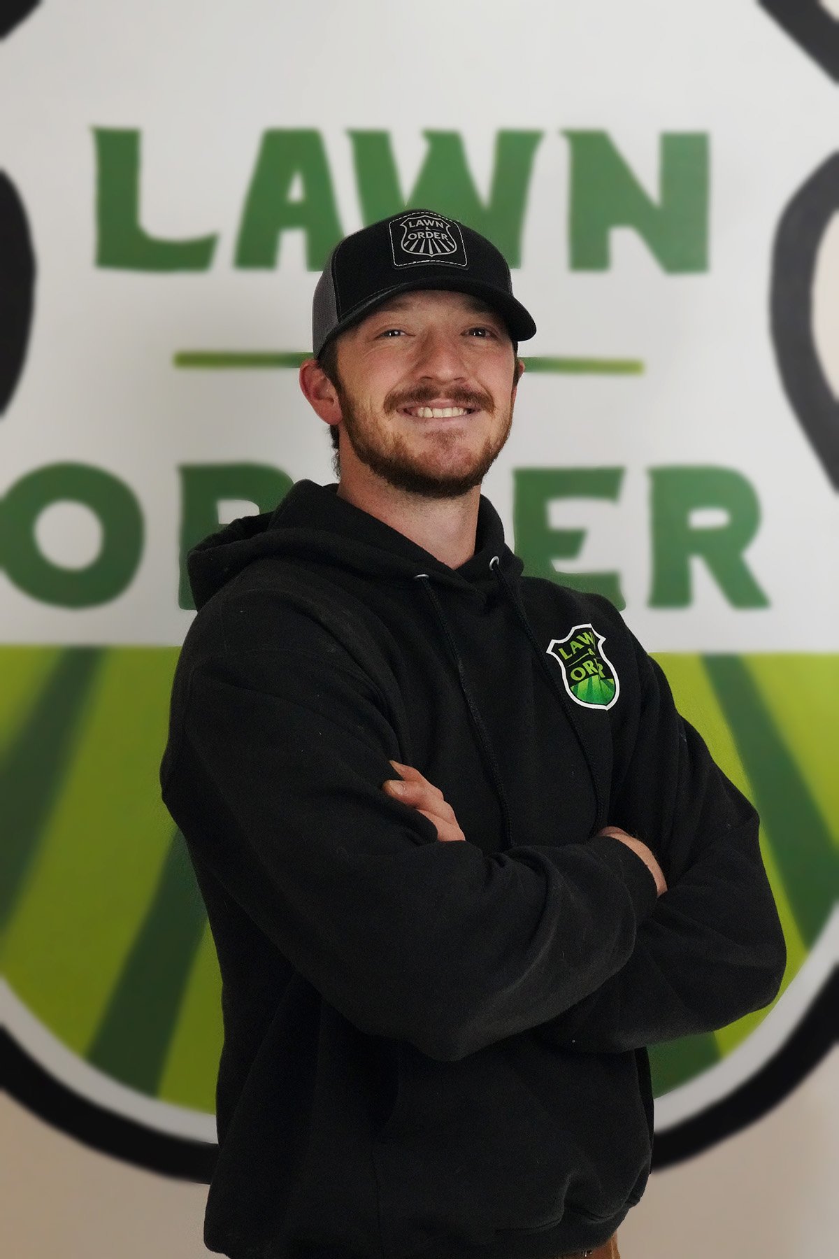 A man smiling with arms crossed, wearing a black hoodie and black cap, standing in front of a large sign that reads 'LAWN' and 'POWER' with a green and white design.