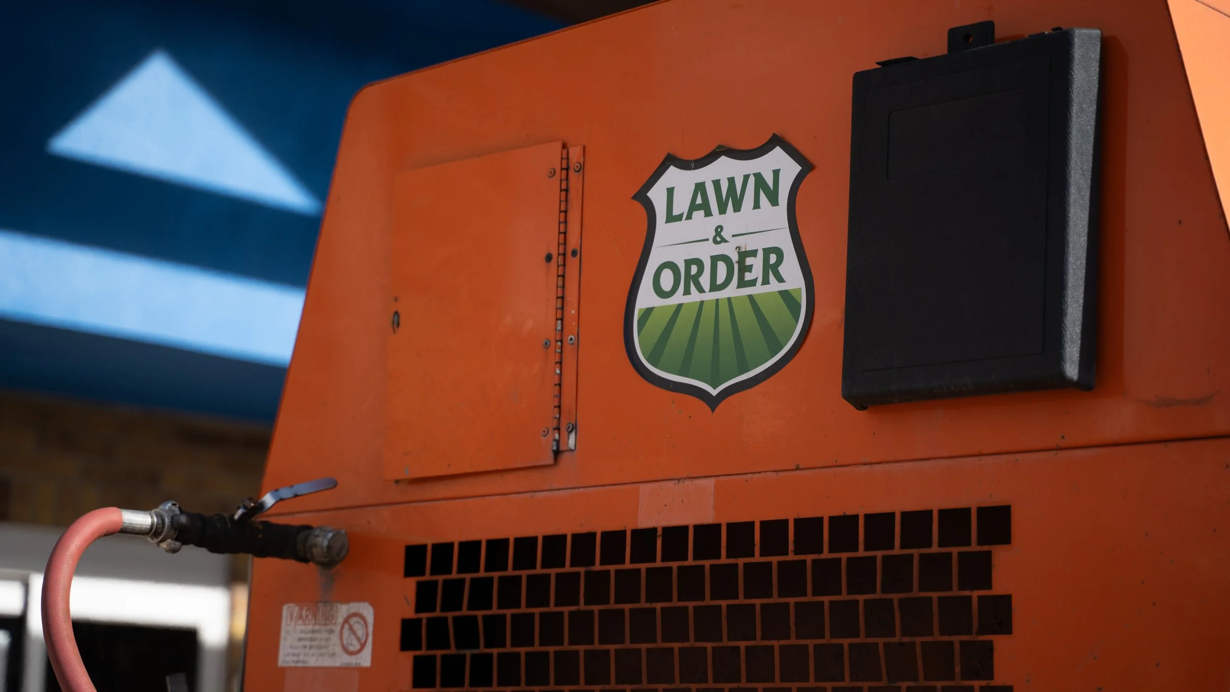 Close-up of an orange lawn equipment with a 'Lawn & Order' sticker on it, featuring a shield-shaped logo with green grass design, and a black rectangular box attached to the right side.