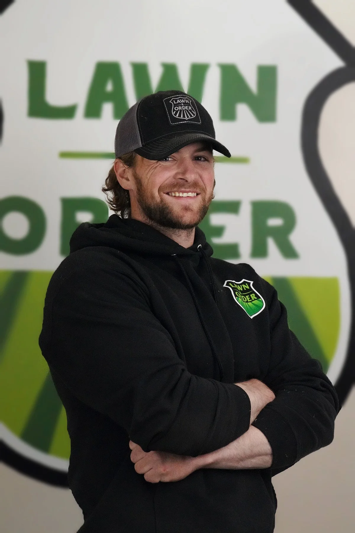 A man with a beard and long hair wearing a black hoodie and a gray and black baseball cap with the logo 'Lawn Order'. He is standing in front of a sign that says 'Lawn Order' with a green and black design, and he is smiling with his arms crossed.
