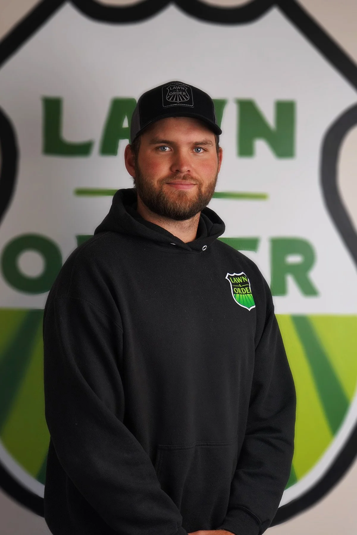 A young man with a beard and blue eyes wearing a black hoodie and a baseball cap with a lawn mower logo, standing in front of a large logo background that reads 'Lawn.'