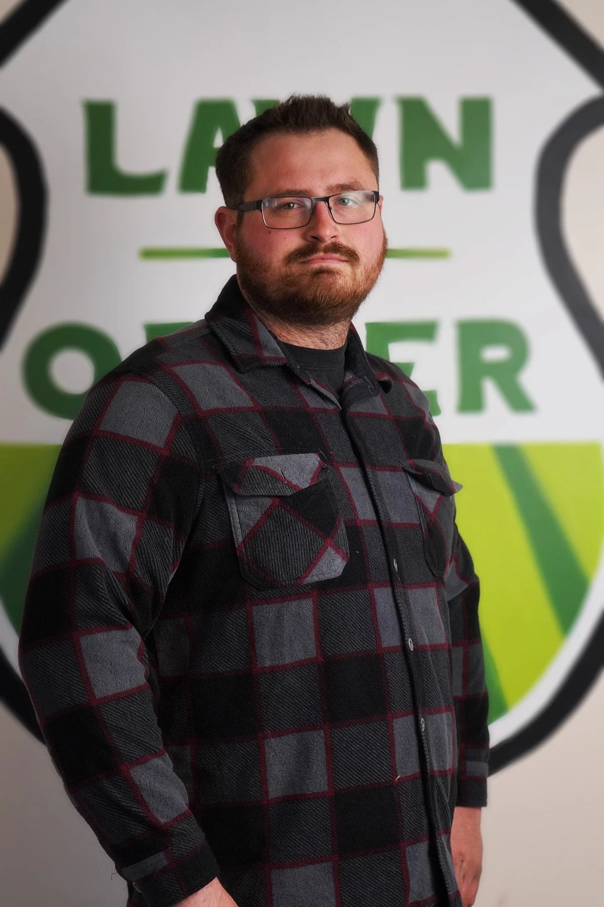 A man with glasses and a beard wearing a black and gray plaid shirt standing in front of a wall with green and white lettering.