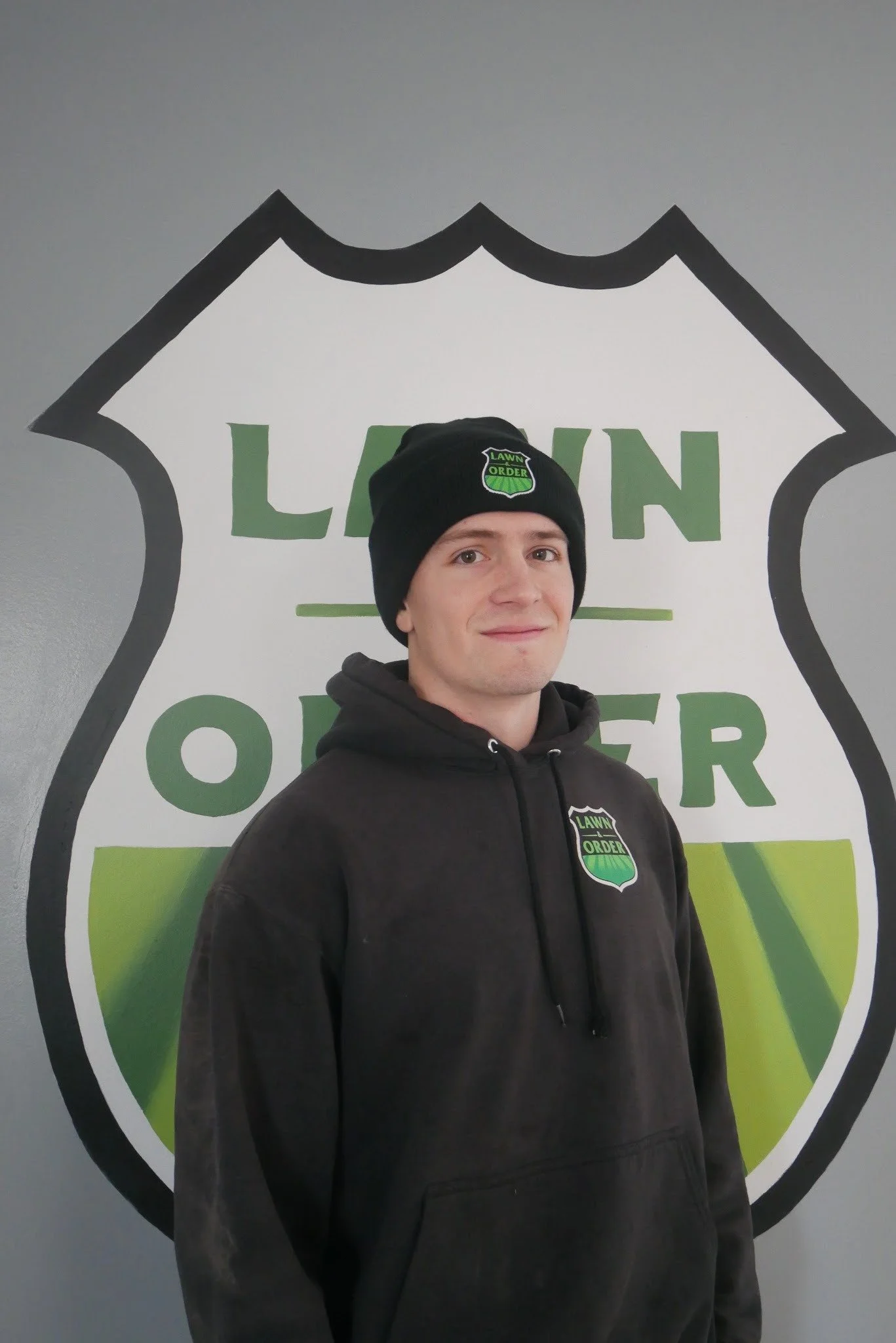 A young man wearing a black hoodie and a black beanie with a "Lawn Order" logo, standing in front of a wall with a large "Lawn Order" logo.