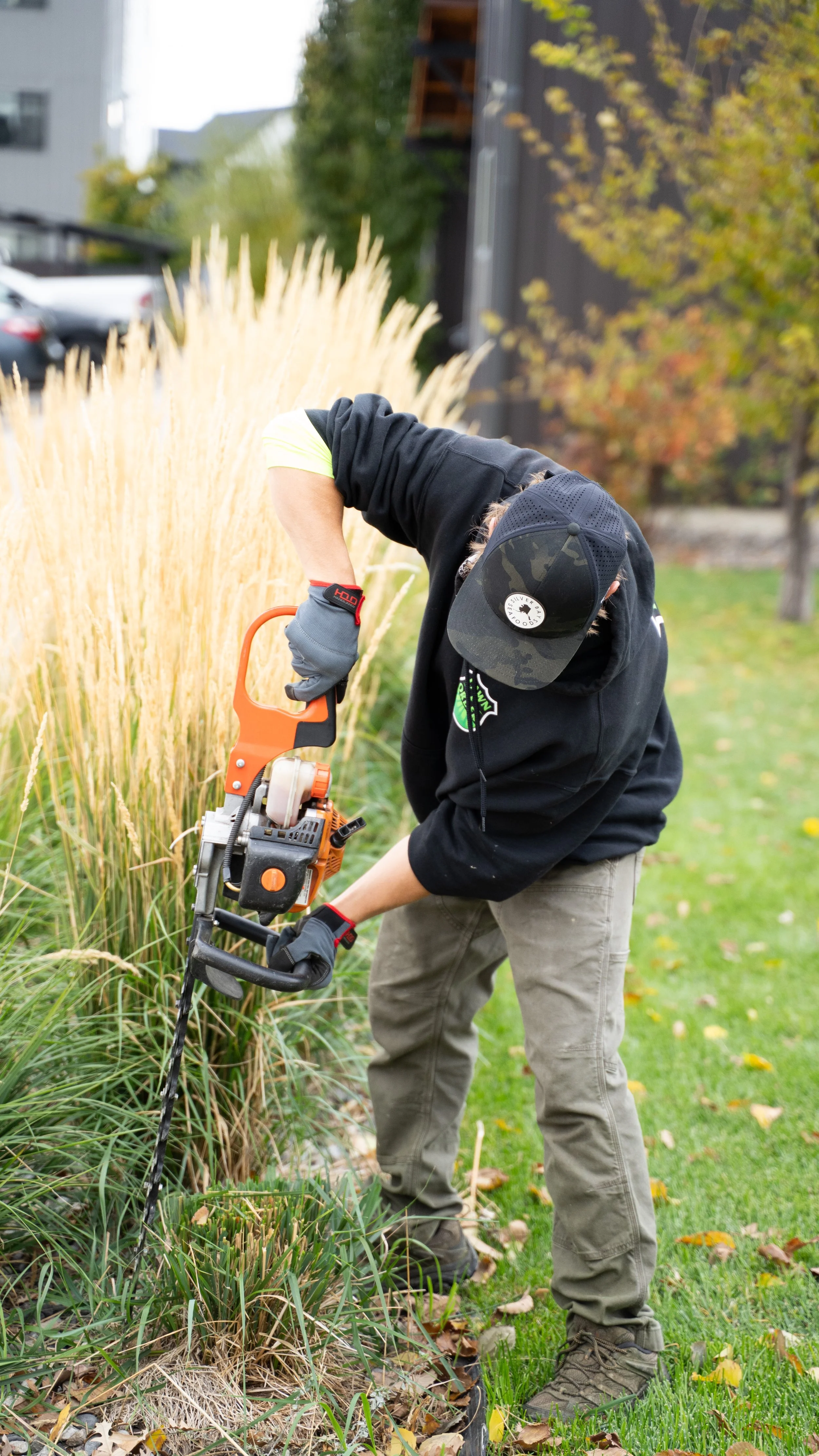 A person using a hedge trimmer to cut tall grass and plants in a garden during fall.