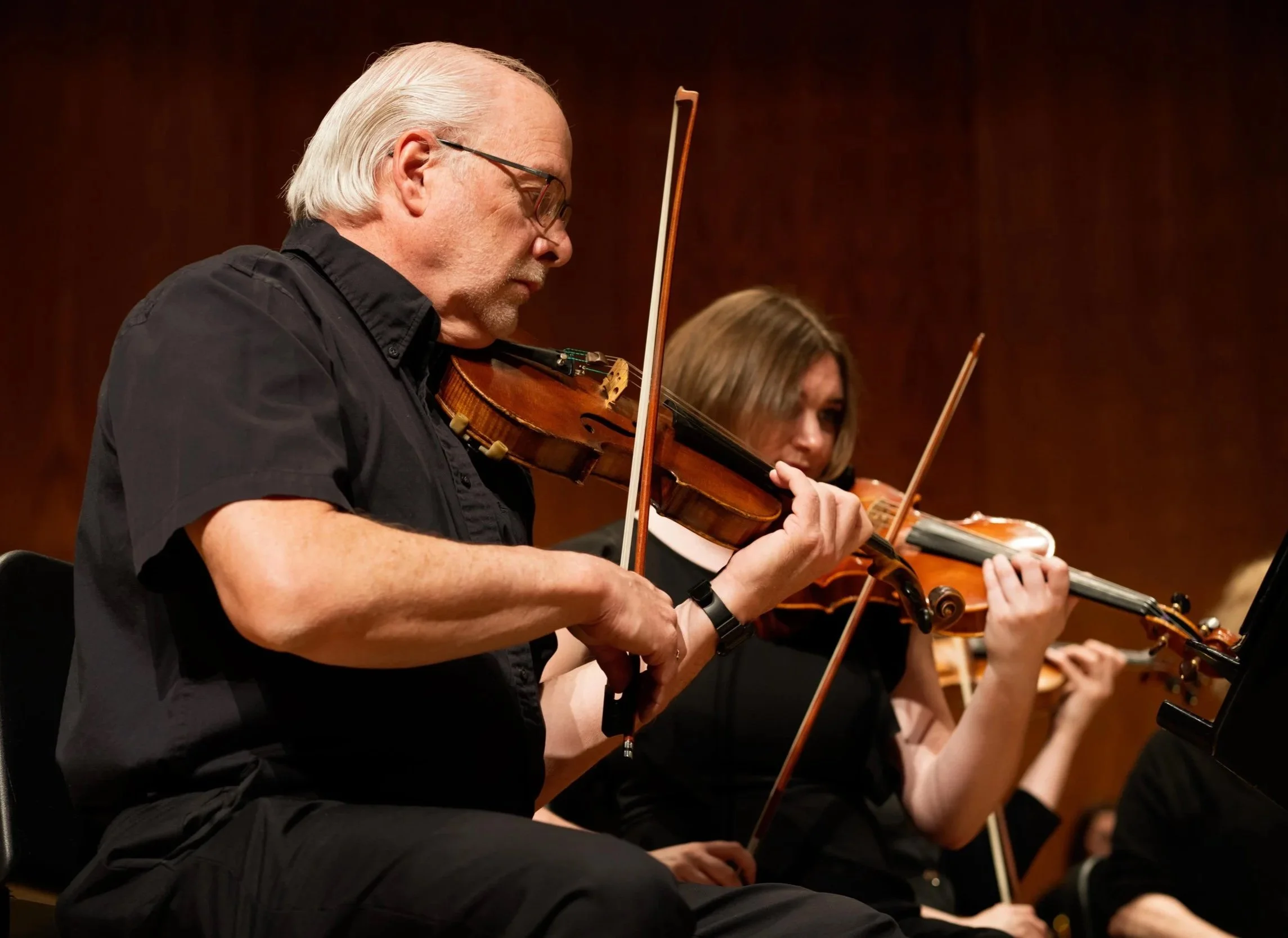 Two musicians playing violins during a performance, with a dark background.