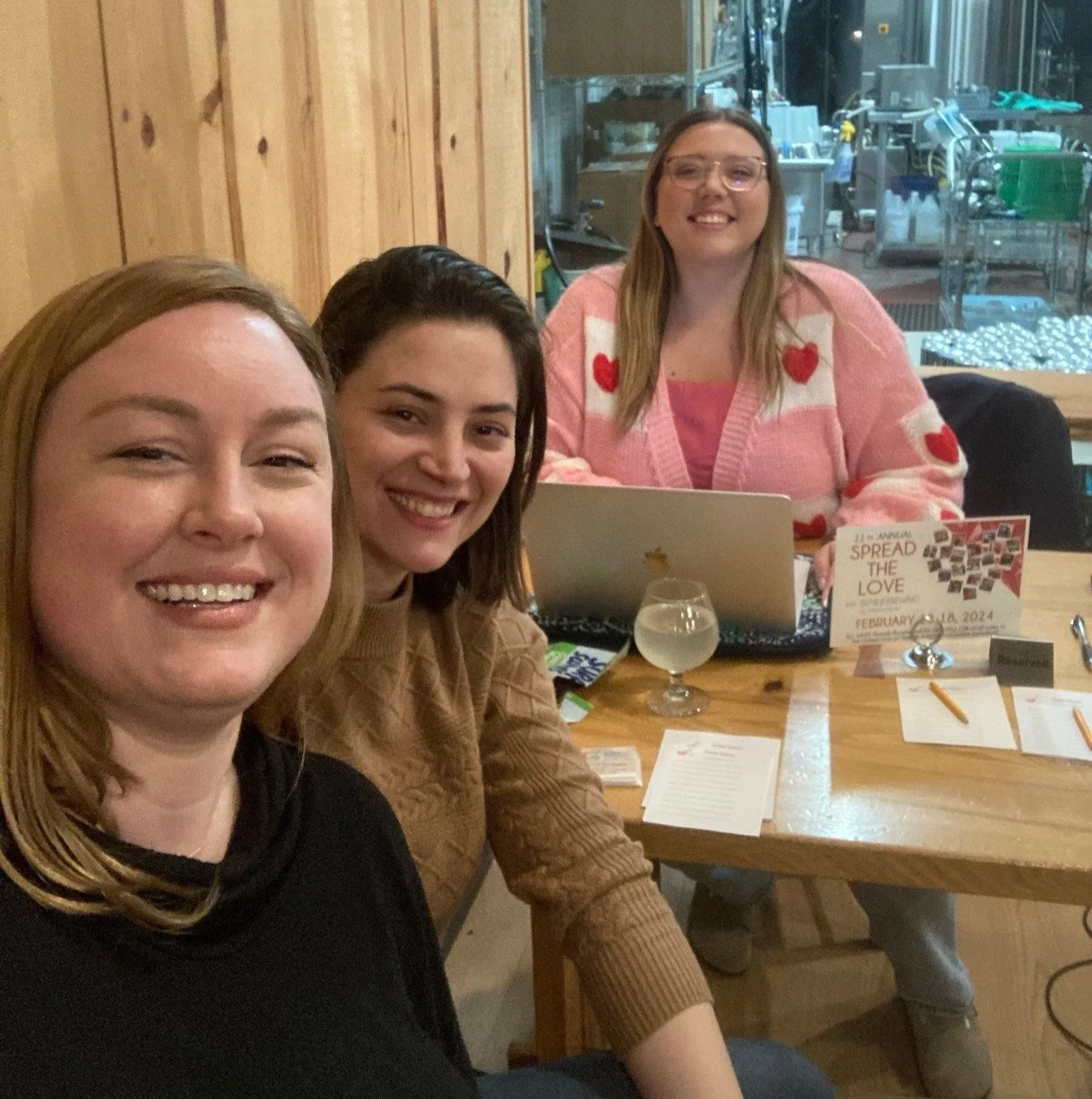 Three women smiling at a table in a restaurant or cafe, with one woman using a laptop. The table has papers, a glass of beverage, and a sign that reads 'Spread the Love' with a date in February 2024. The background shows a window and kitchen area.