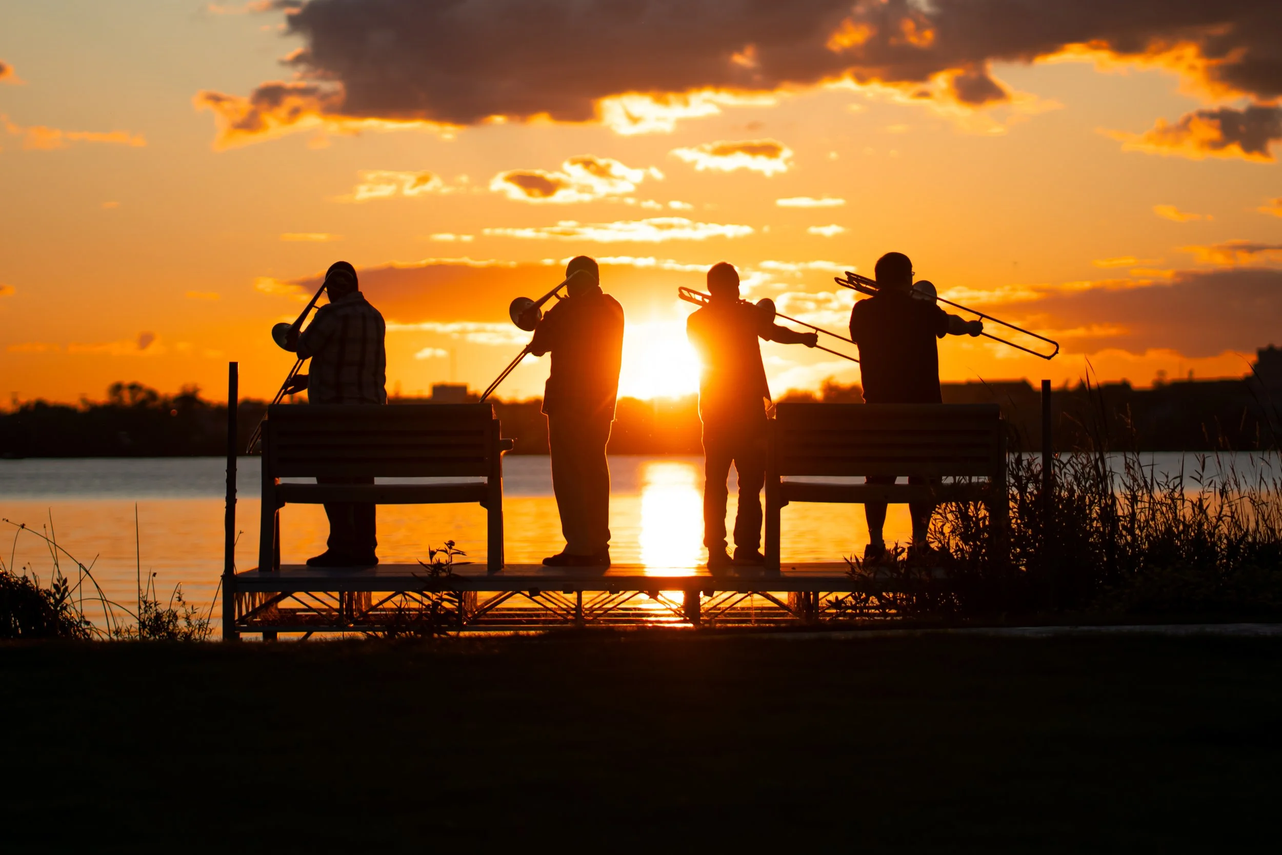 Four people sitting on a pier by the water during sunset, each playing a trombone silhouetted against the colorful sky.