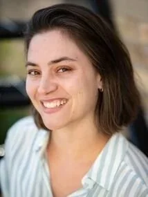Young woman smiling outdoors with dark hair, wearing a light-colored striped shirt.