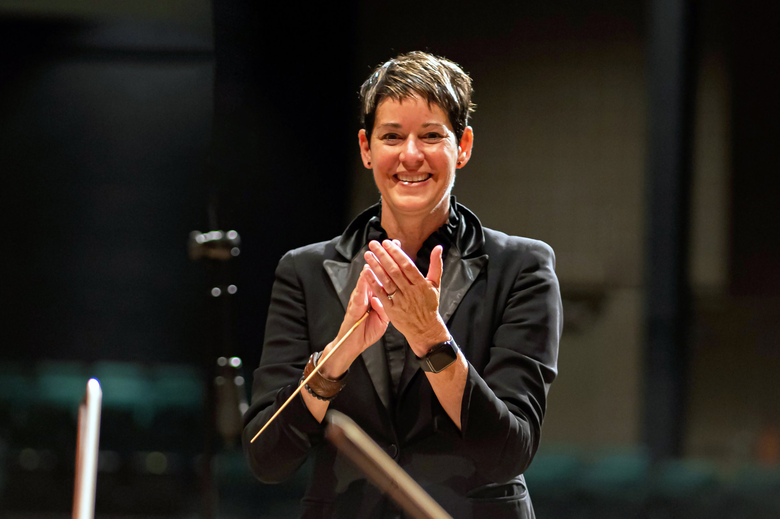 A woman with short dark hair smiling and clapping in a dark room, dressed in a black jacket and wearing a smartwatch.