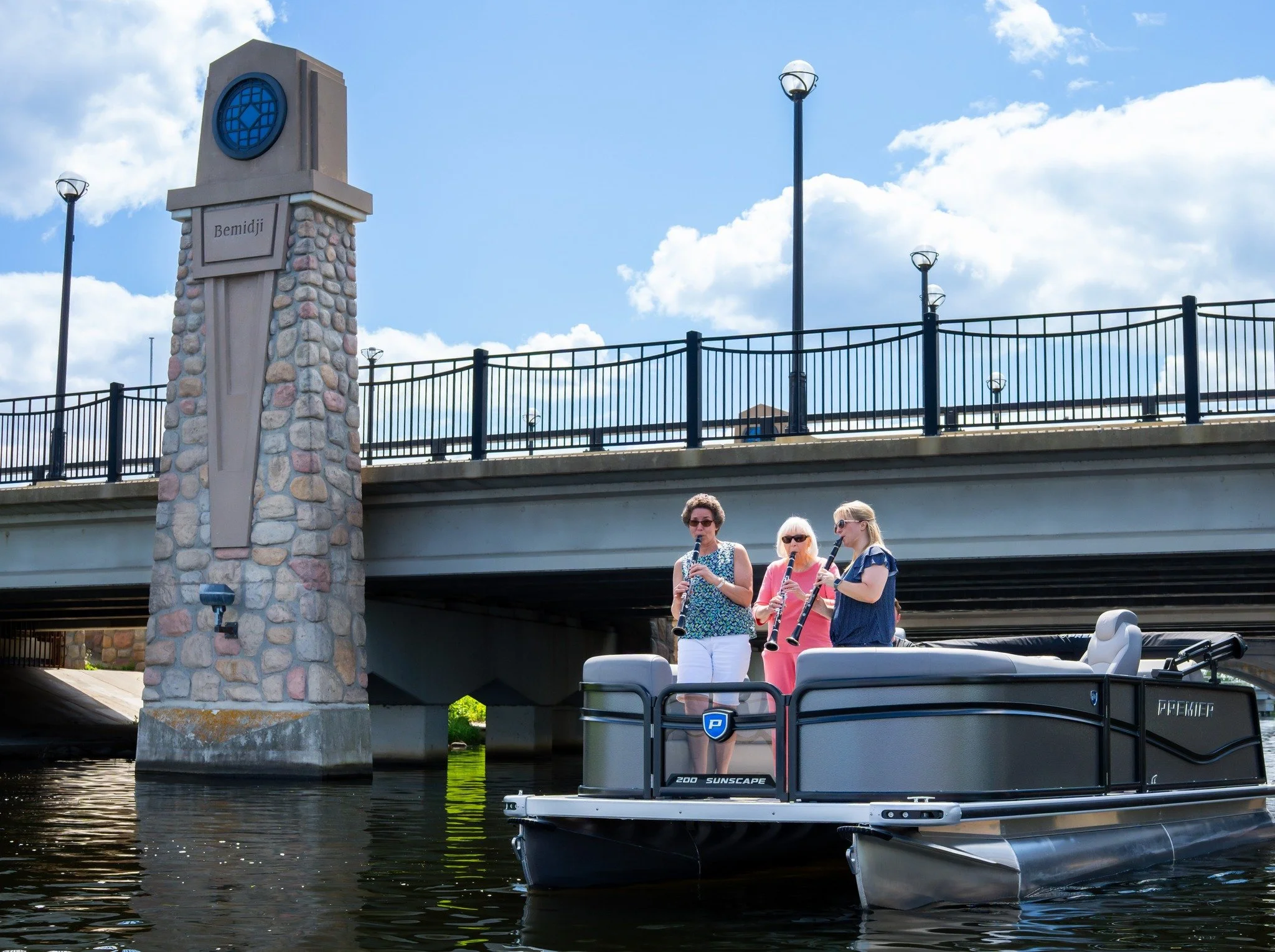 Three women stand on a pontoon boat on a river, playing wind instruments near a stone bridge with lamps and a clock tower that has the word 'Bemidji' on it, under a partly cloudy sky.